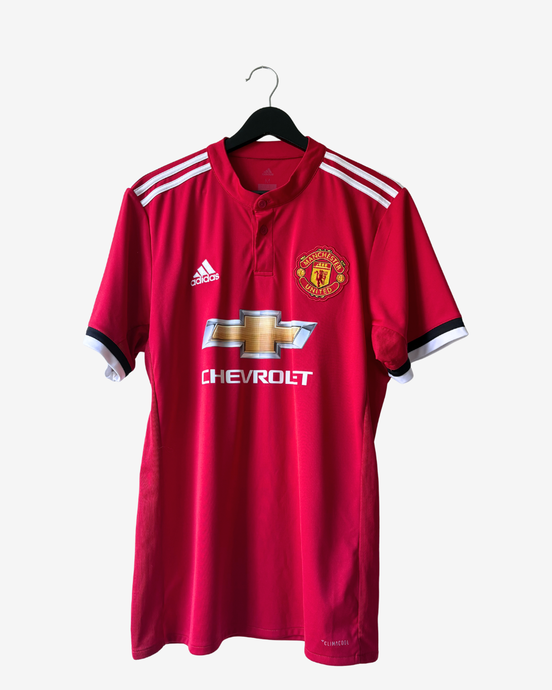 A red Manchester United short-sleeve soccer jersey by Adidas, featuring the club's crest, Chevrolet sponsor logo, and white shoulder stripes, hanging on a black hanger against a light gray background.