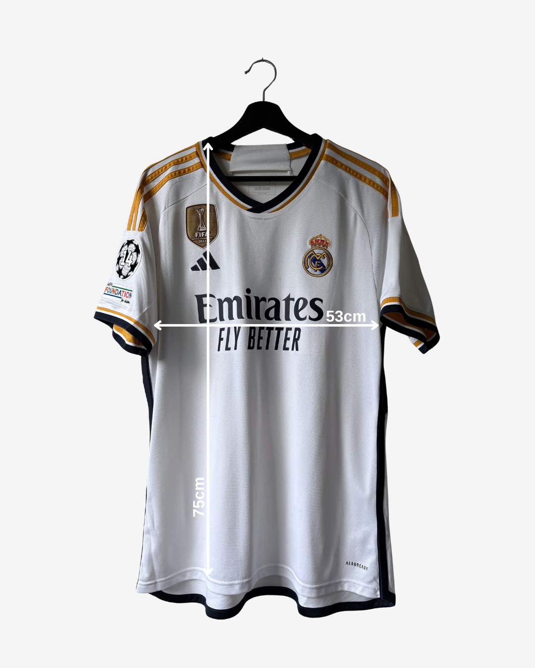 A white Real Madrid soccer jersey with gold and black accents, hanging on a black hanger against a light gray background. The jersey features the Emirates Fly Better sponsor logo, the Adidas logo, the Real Madrid crest, and UEFA Champions League patches on the sleeve.