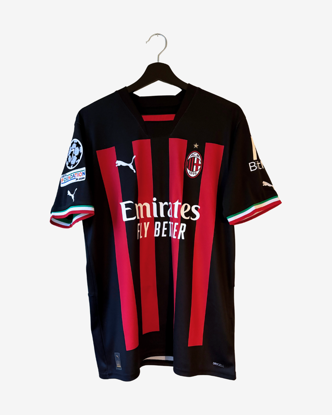 A black and red striped AC Milan soccer jersey by Puma, featuring the Emirates Fly Bezer sponsor logo, hanging on a black hanger against a light gray background.