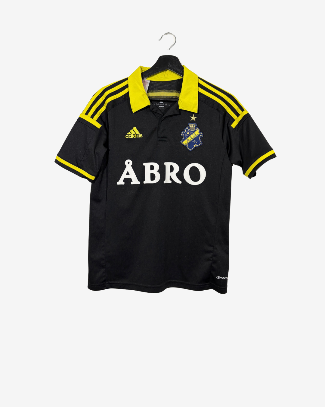 A black Adidas soccer jersey with yellow collar and shoulder stripes, featuring the white 'ÅBRO' sponsor logo on the front, hanging on a black hanger against a light gray background.