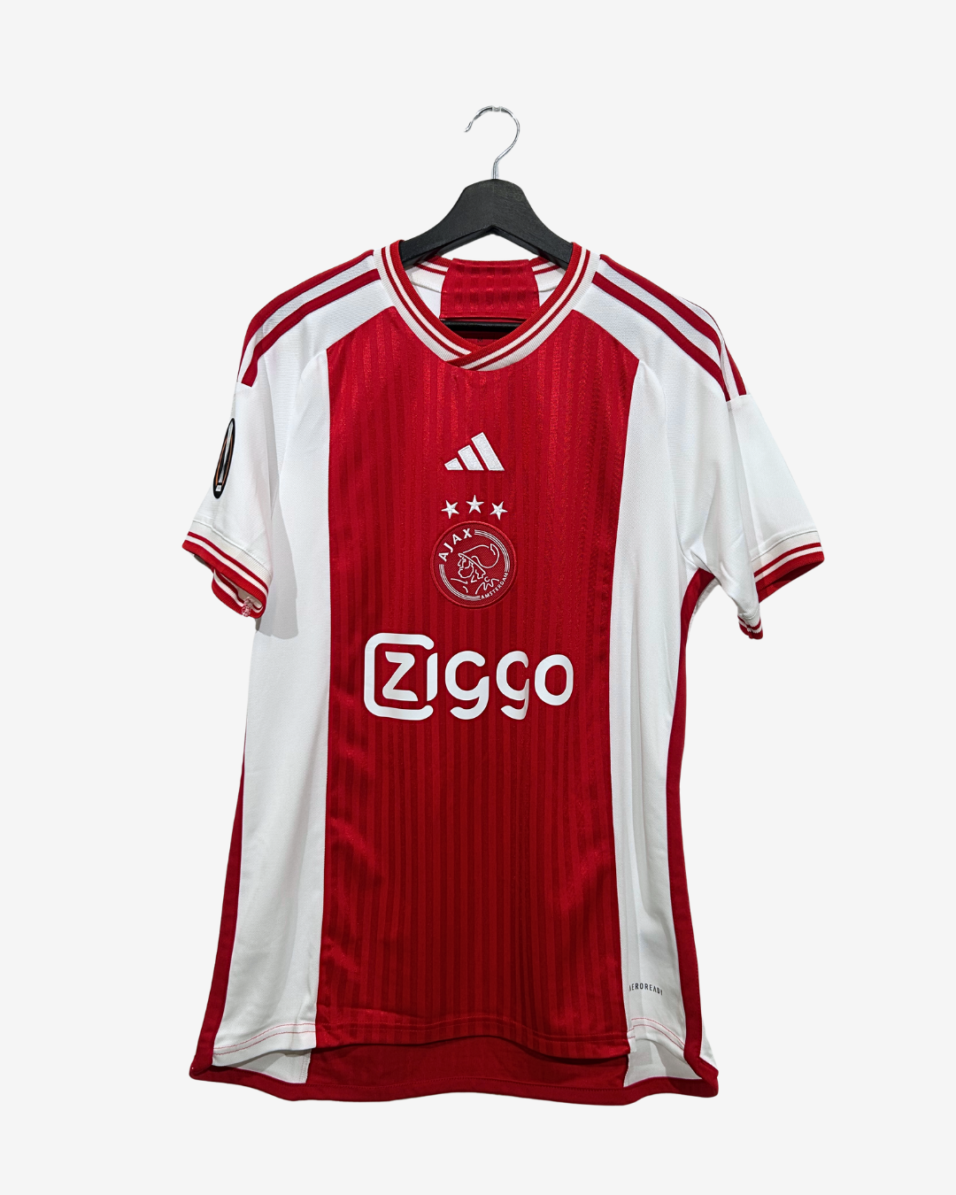 A red and white Ajax Amsterdam soccer jersey by Adidas, hanging on a black hanger against a light gray background. The jersey features the Ziggo sponsor logo on the front.