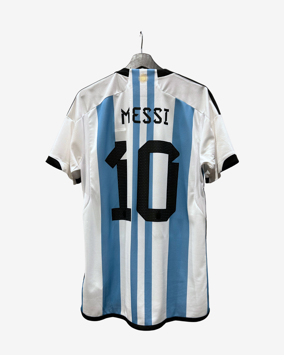 Back view of an Argentina national football team home jersey featuring light blue and white vertical stripes, with 'MESSI' and the number '10' printed in black on the back. The jersey is displayed on a hanger against a light gray background.