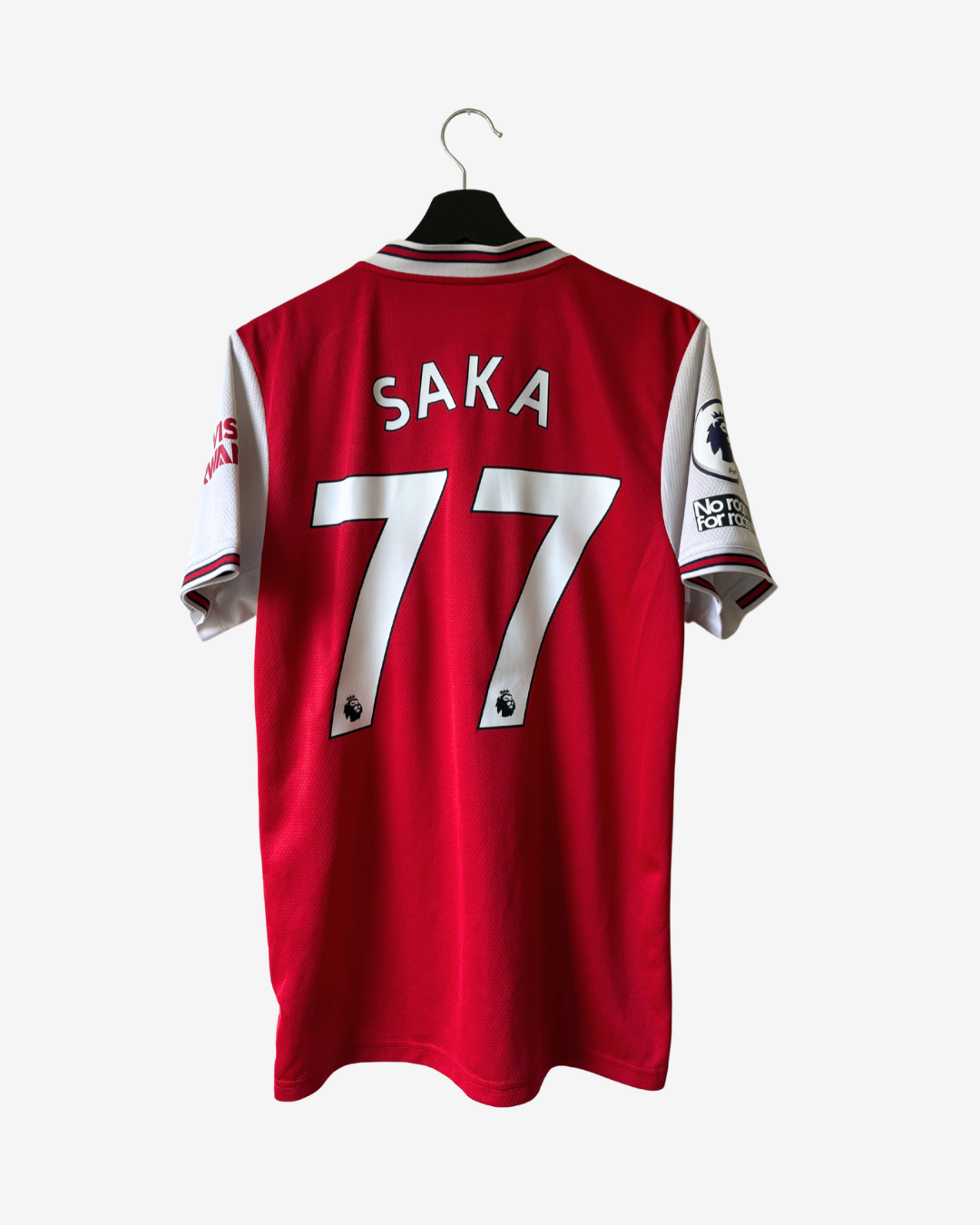A red short-sleeve soccer jersey hanging on a black hanger against a light gray background. The back of the jersey features the name 'SAKA' and the number '77' in white, with white shoulder panels and sleeve details.