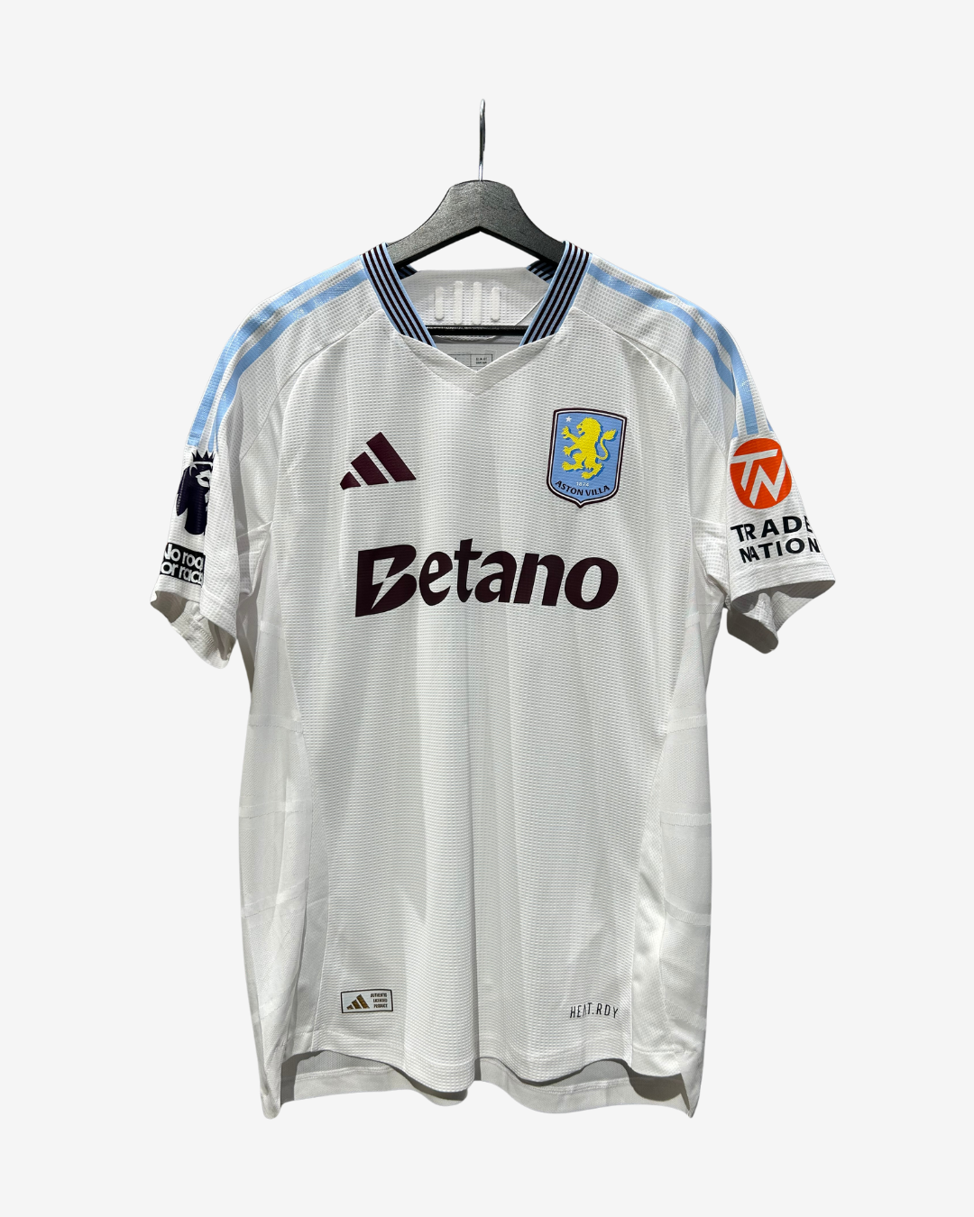 A white Adidas Aston Villa football jersey with light blue shoulder stripes, hanging on a black hanger against a light gray background. The jersey features the Betano sponsor logo across the chest, the club crest, and the Adidas logo.