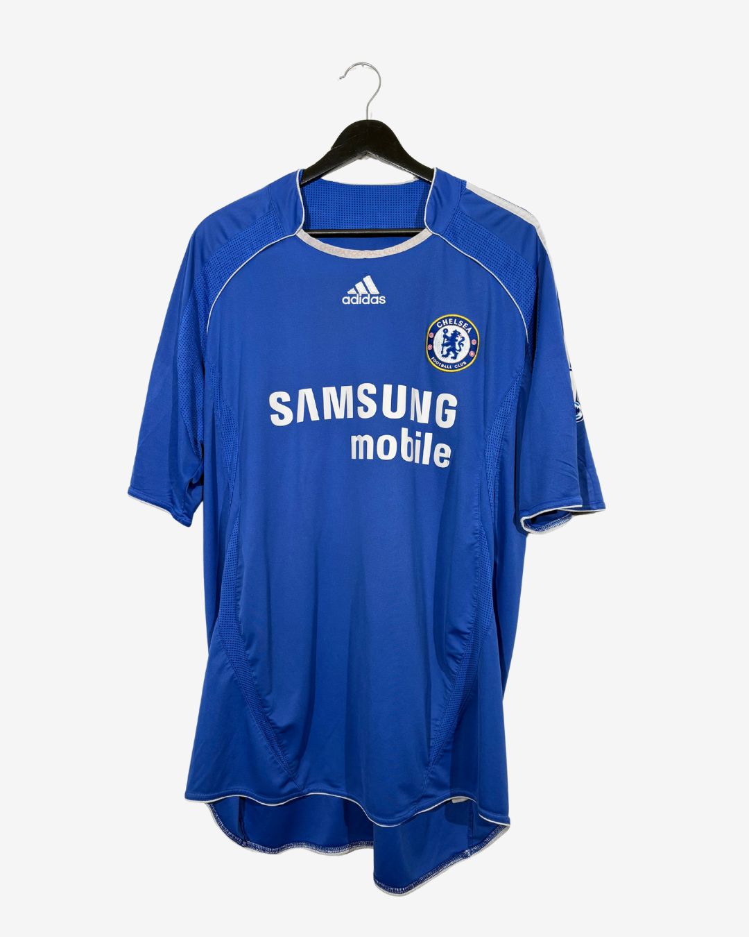 A blue Chelsea football jersey with white trim, featuring the Samsung motile sponsor logo and the Adidas logo, hanging on a black hanger against a light gray background.