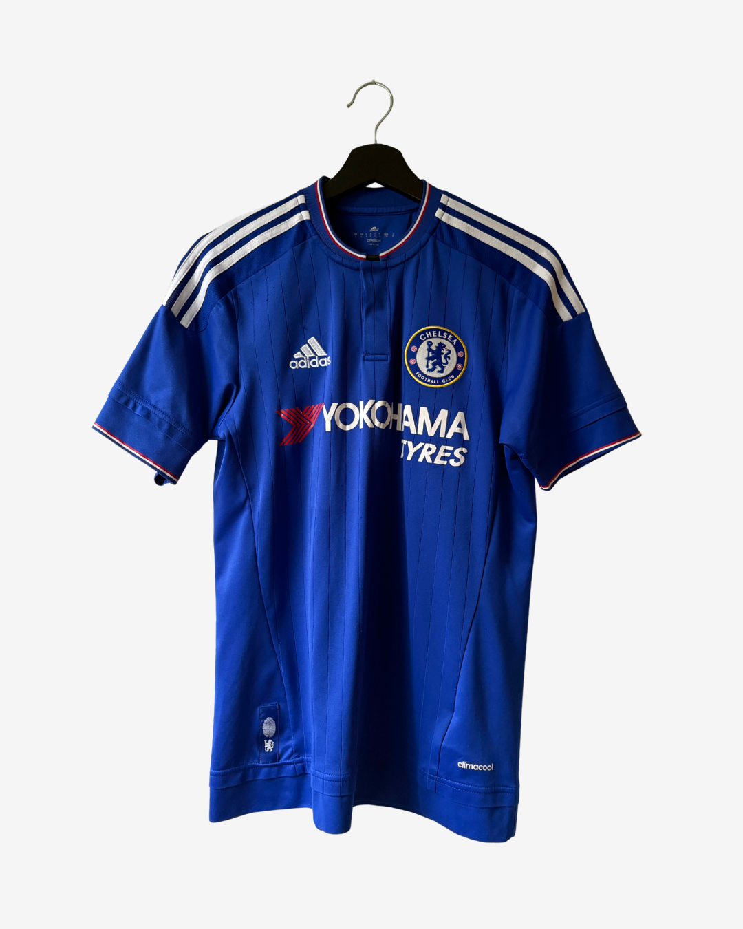 A blue Chelsea football jersey with white shoulder stripes, hanging on a black hanger against a light gray background. The jersey features the Adidas logo, Chelsea crest, and Yokohama Tyres sponsor logo.