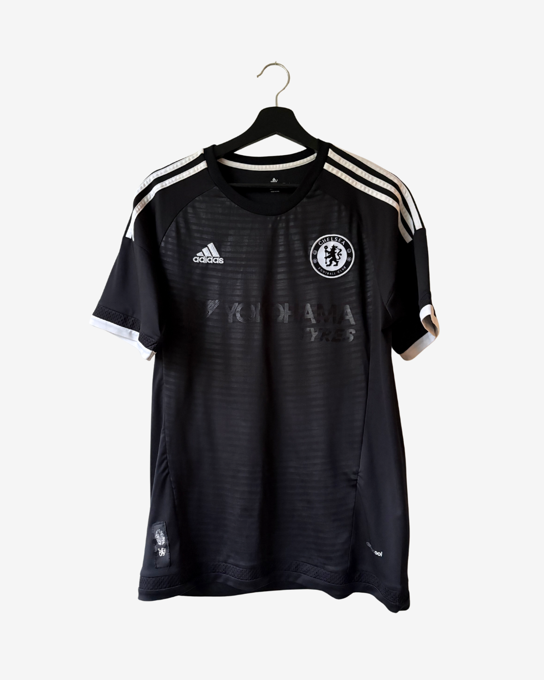 A black Chelsea FC home soccer jersey by Adidas, featuring white shoulder stripes, the club crest on the chest, hanging on a black hanger against a light gray background.