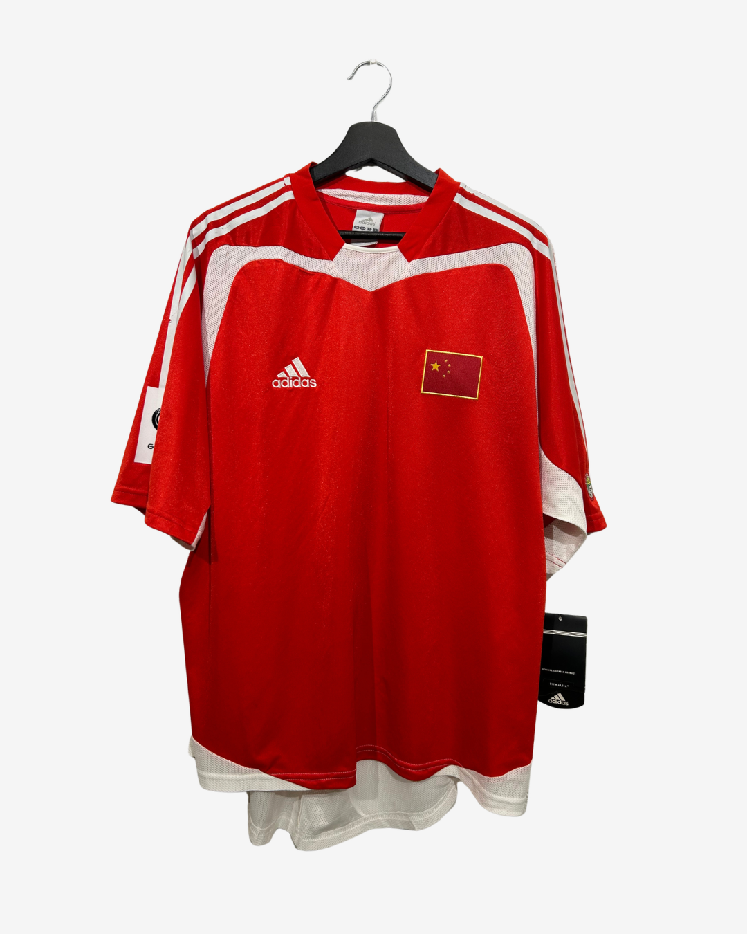 A red Adidas soccer jersey with white shoulder stripes and a white hem, featuring a Chinese flag patch on the chest, hanging on a black hanger against a light gray background.