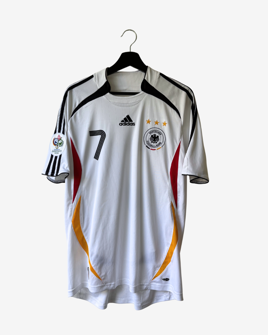 A white Adidas Germany national football team jersey with black, red, and yellow accents, featuring the number 7 on the right chest and three gold stars, hanging on a black hanger against a light gray background.
