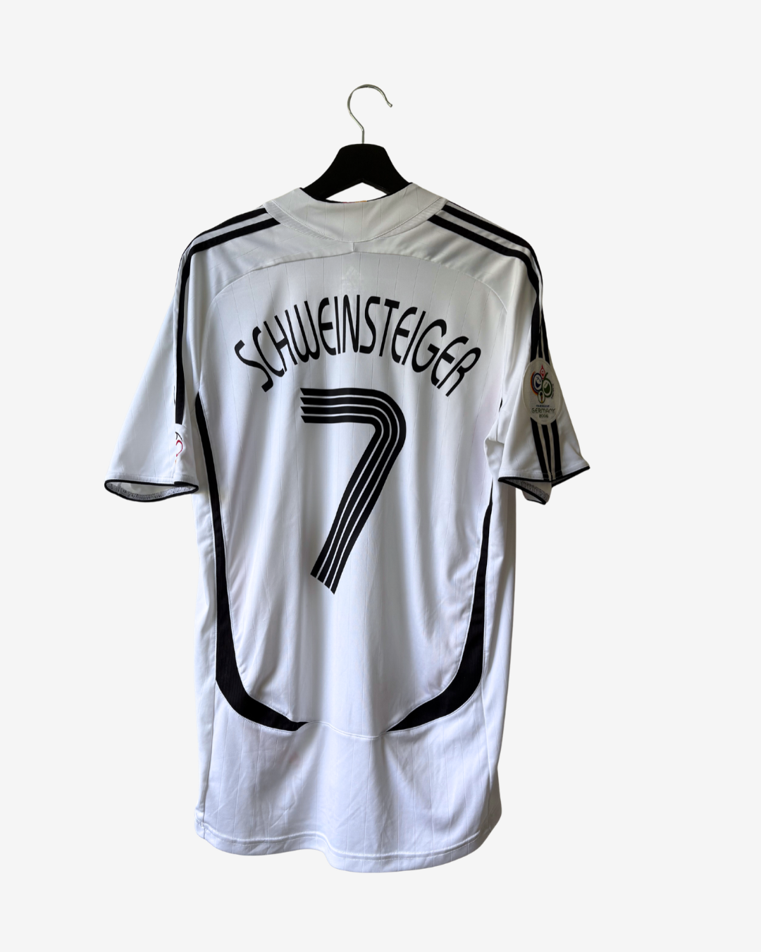 A white Germany national football team home jersey with 'SCHWEINSTEIGER' and the number '7' printed on the back, hanging on a black hanger against a light gray background.
