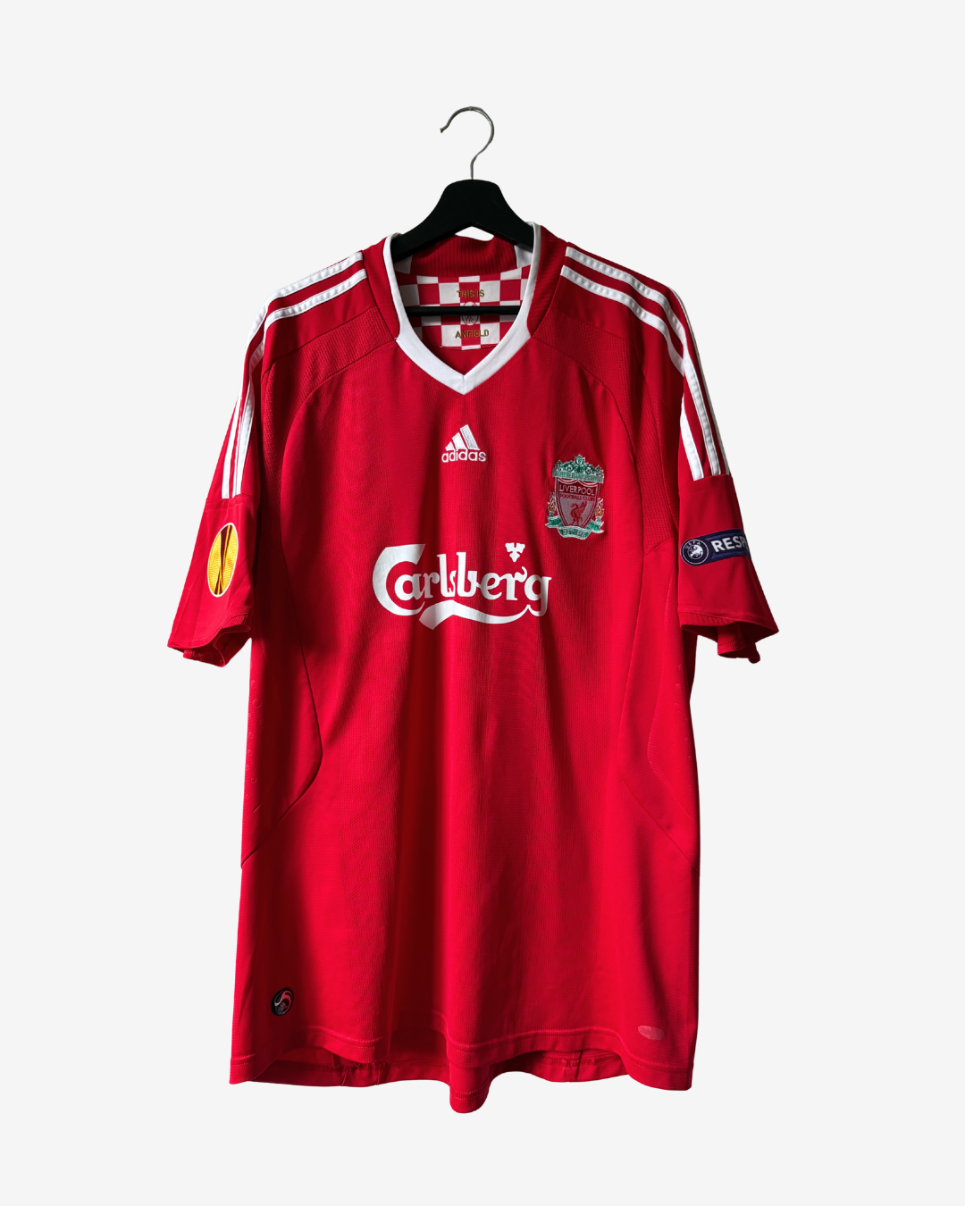 A red Adidas Liverpool football jersey hanging on a black hanger against a light gray background. The jersey features the white Carbery sponsor logo on the chest, the Adidas logo, the Liverpool club crest, and white stripes on the shoulders and sleeves.