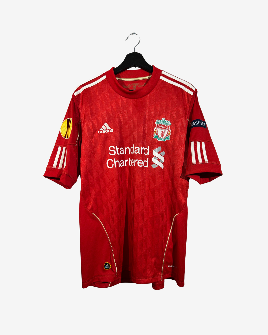 A red Adidas Liverpool football jersey hanging on a black hanger against a light gray background. The jersey features white stripes on the shoulders and sleeves, the Liverpool club crest on the chest, the Standard Chartered sponsor logo, and a UEFA Champions League patch on the sleeve.