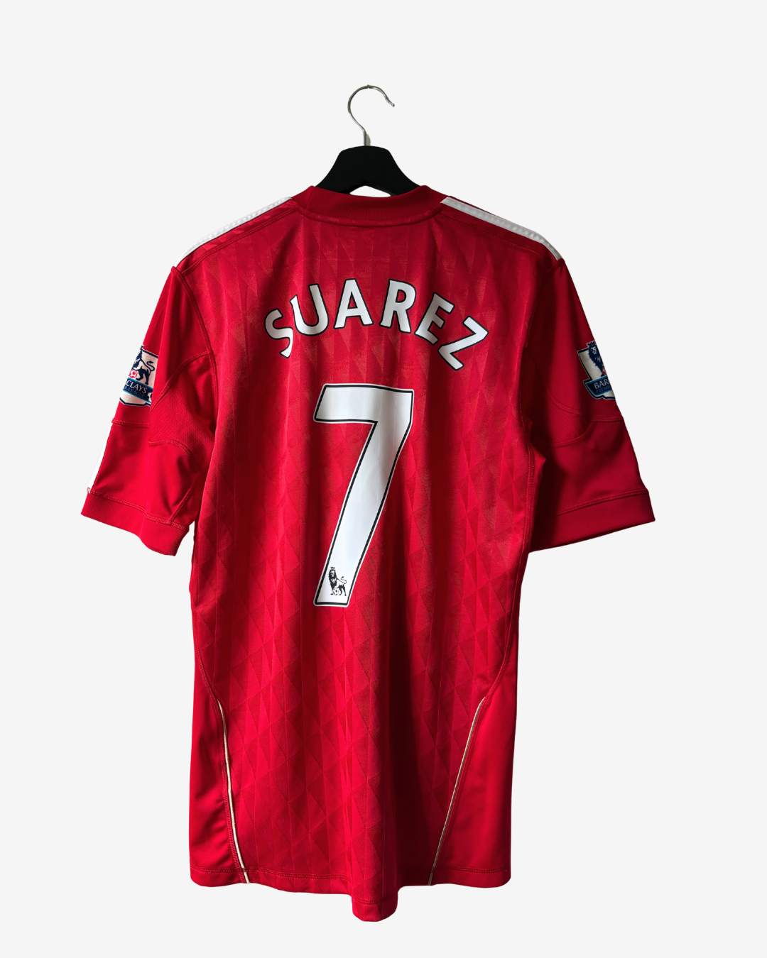 A red short-sleeve soccer jersey hanging on a black hanger against a light gray background. The back of the jersey features the name 'SUAREZ' and the number '7' in white with black outlines.