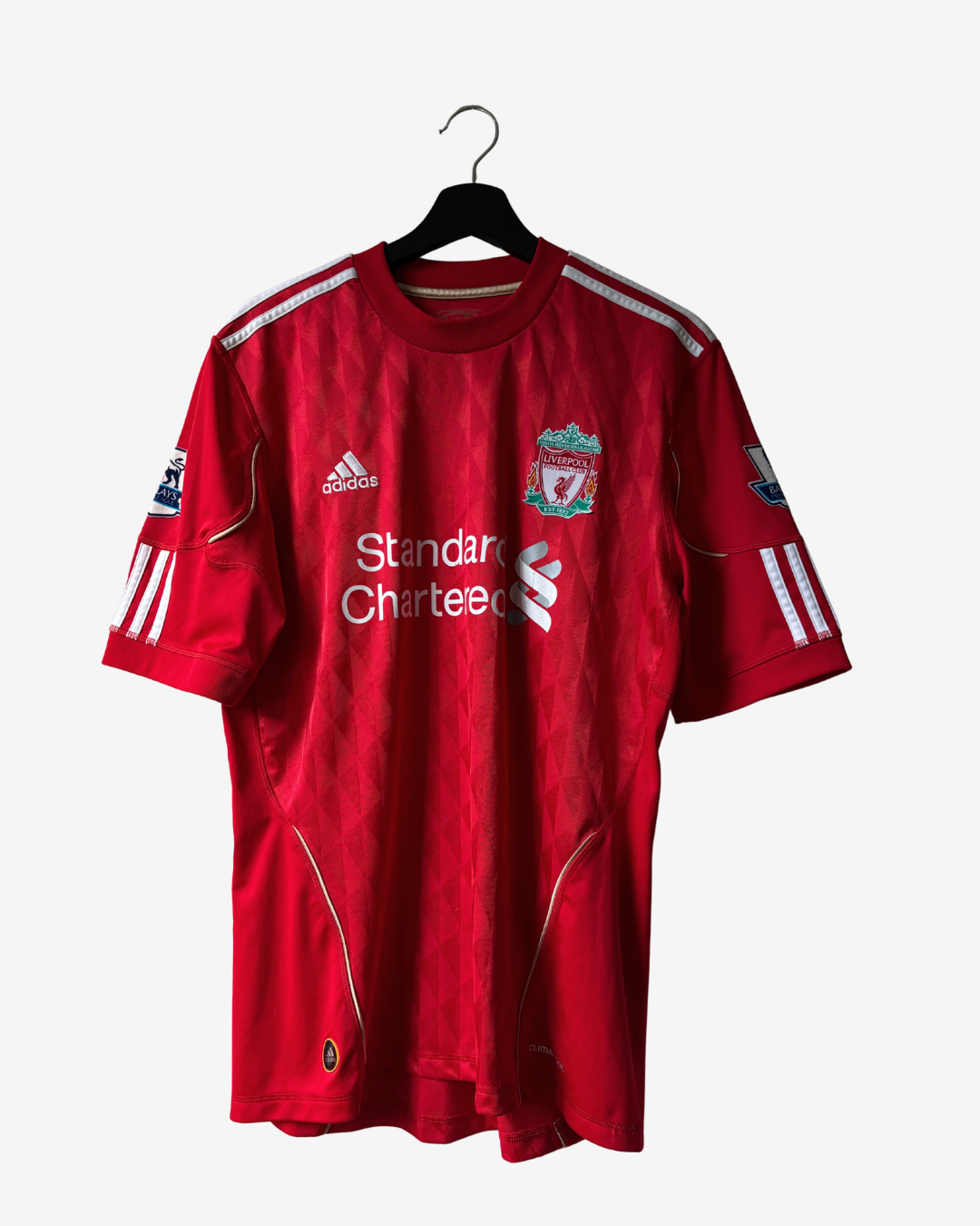 A red Adidas Liverpool football jersey hanging on a black hanger against a light gray background. The jersey features the Liverpool club crest on the chest, the white Standard Chartered sponsor logo, and white Adidas branding.