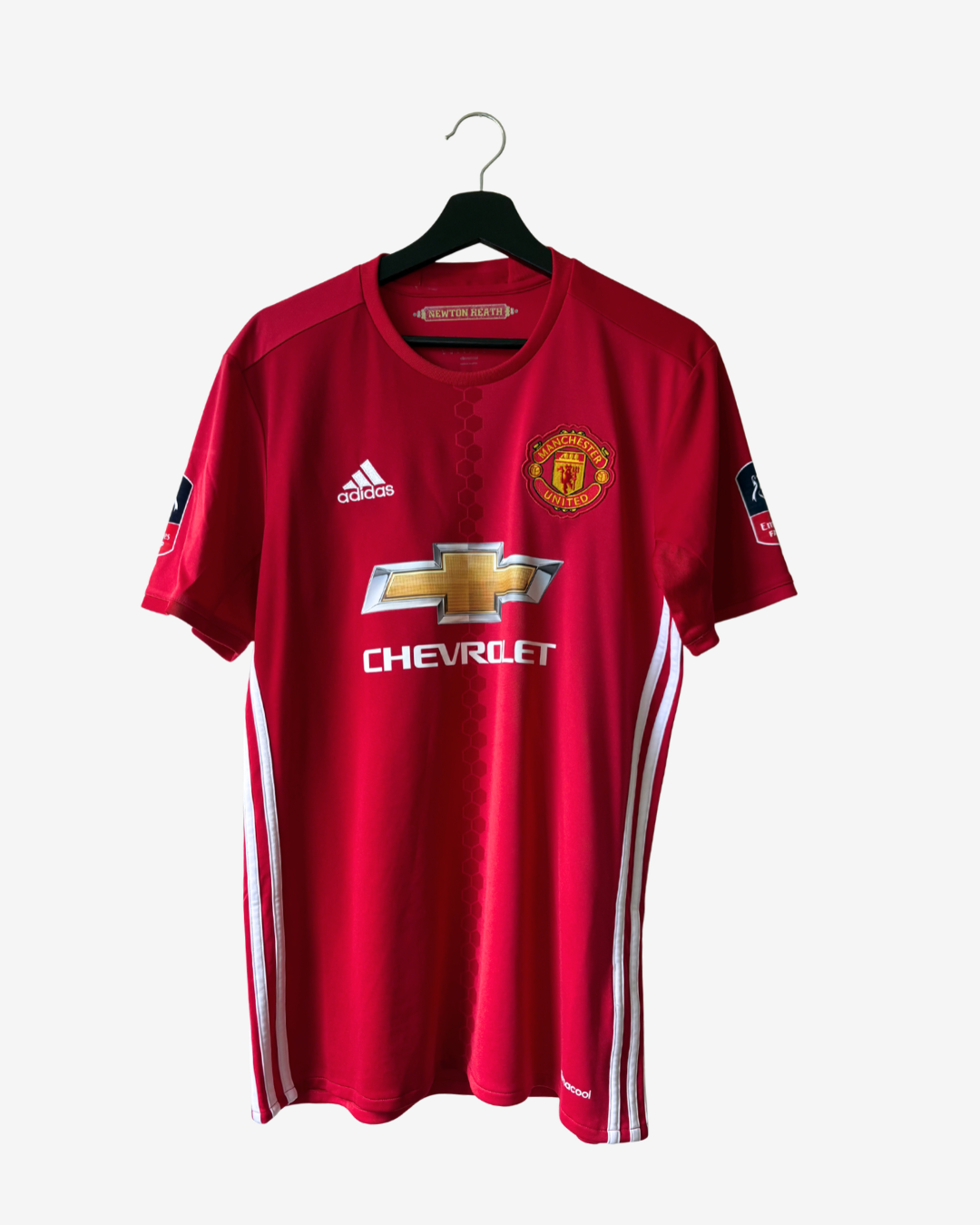 A red Manchester United soccer jersey by Adidas, hanging on a black hanger against a light gray background. The jersey features the Manchester United crest on the left chest, the Adidas logo on the right chest, and the Chevrolet sponsor logo across the center.