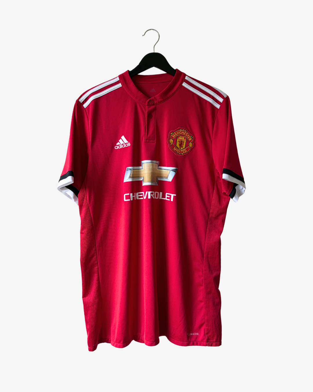 A red Manchester United soccer jersey by Adidas, featuring the club's crest, Chevrolet sponsor logo, and white shoulder stripes, hanging on a black hanger against a light gray background.