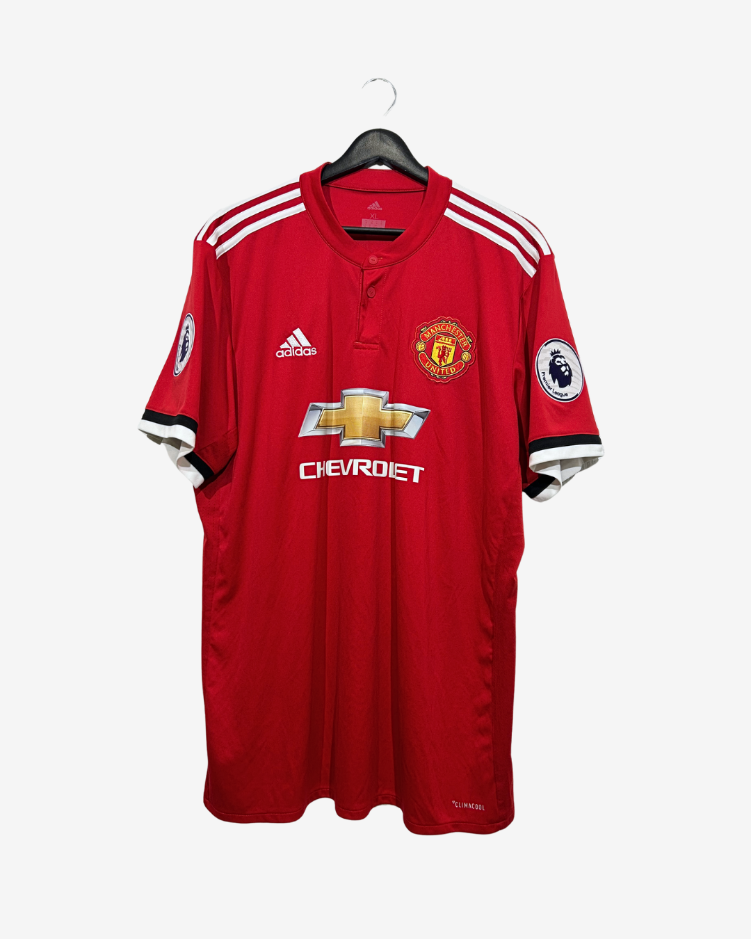 A red Manchester United football jersey by Adidas, featuring the club's crest, Chevrolet sponsor logo, and Premier League patches, hanging on a black hanger against a light gray background.