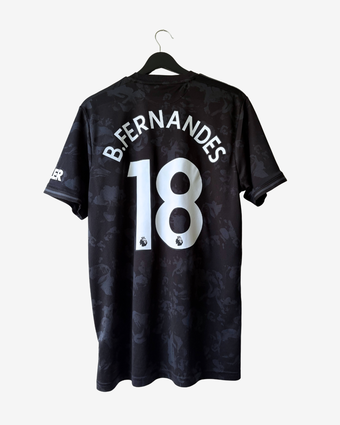 Black Manchester United short-sleeve soccer jersey with 'B. FERNANDES' and number '18' printed in white on the back, hanging on a black hanger against a light gray background.