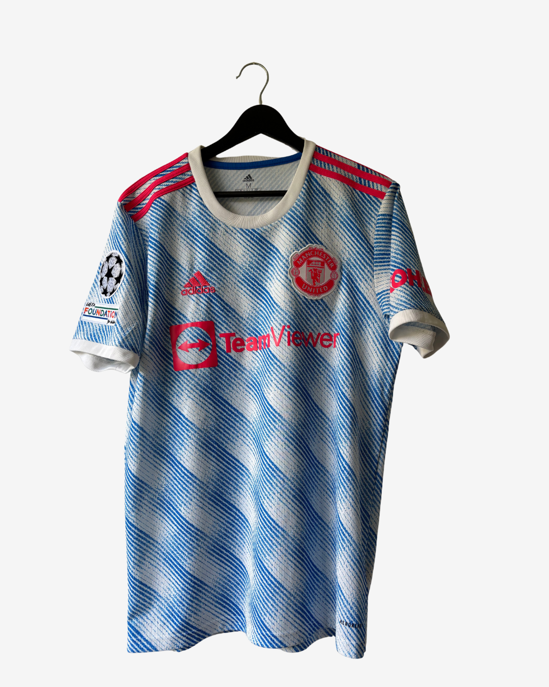 A Manchester United home soccer jersey by Adidas, featuring a light blue and white wavy striped pattern with red accents on the shoulders and red printed logos, hanging on a black hanger against a light gray background.