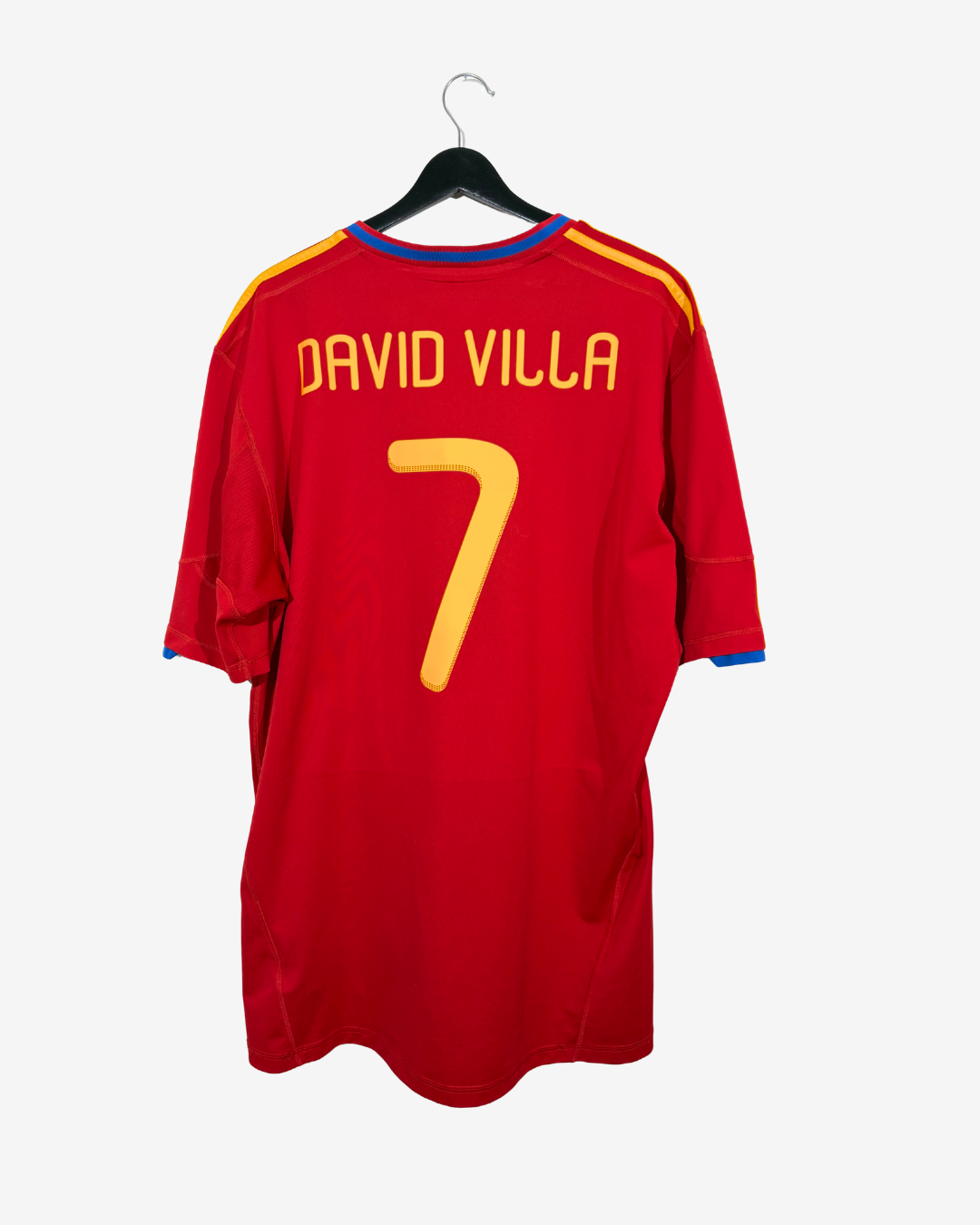 A red short-sleeve soccer jersey hanging on a black hanger against a light gray background. The back of the shirt features 'DAVID VILLA' and the number '7' in yellow, with yellow and blue trim details on the shoulders and collar.