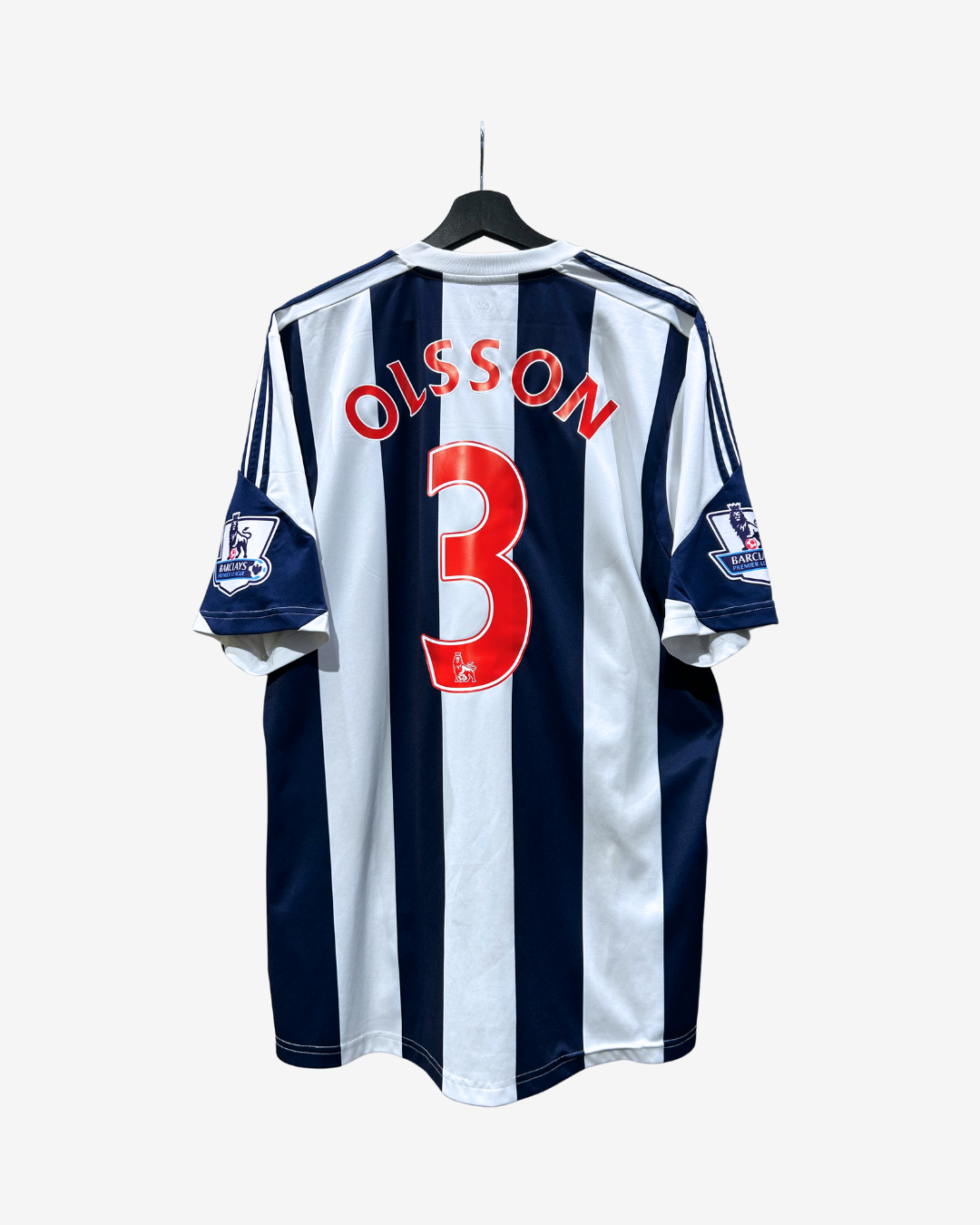 A vintage West Ham United football jersey with navy and white vertical stripes, featuring 'OLSSON' and the number '3' in red on the back. The jersey is hanging on a black hanger against a light gray background.