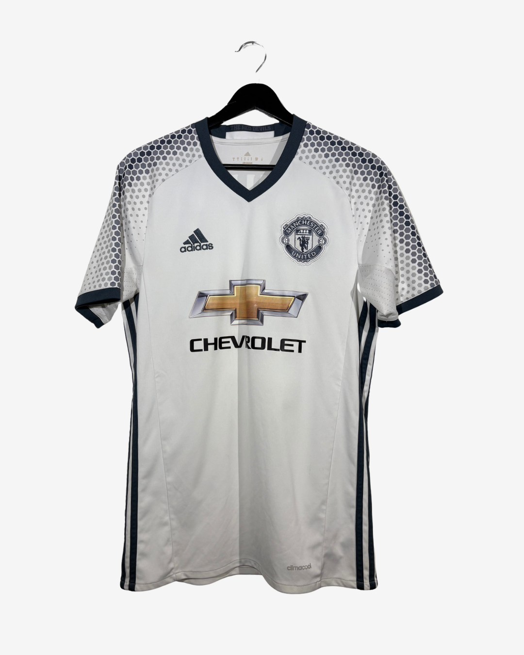 A white Adidas Manchester United soccer jersey with black and gray geometric patterns on the shoulders and sleeves, featuring the gold Chevrolet logo and Manchester United crest on the front, hanging on a black hanger against a light gray background.
