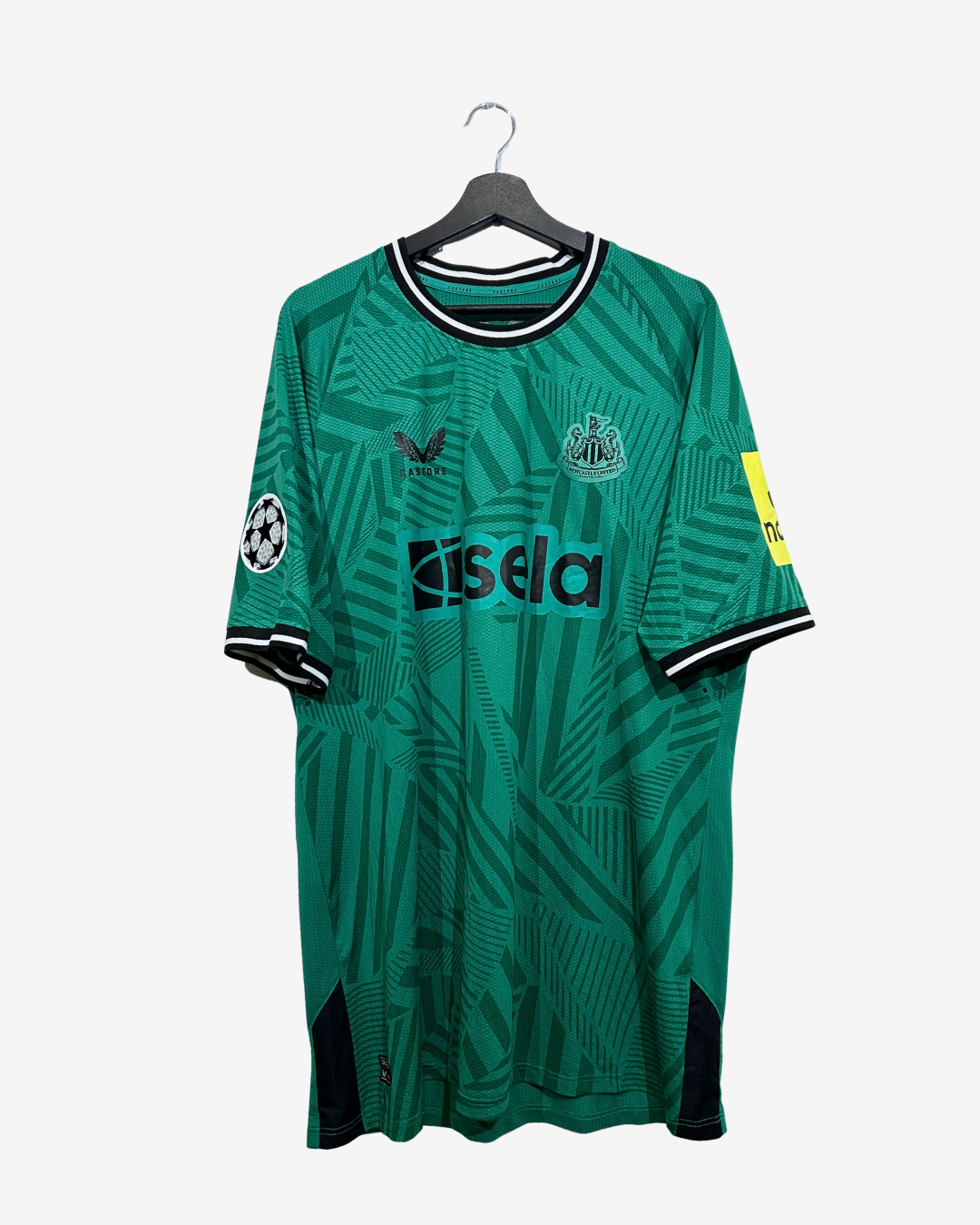 A green and black patterned soccer jersey hanging on a black hanger against a light gray background. The jersey features the Newcastle United crest, a black Vendi logo, a black isela sponsor logo, a UEFA Champions League patch on the right sleeve, and a yellow sleeve patch on the left.