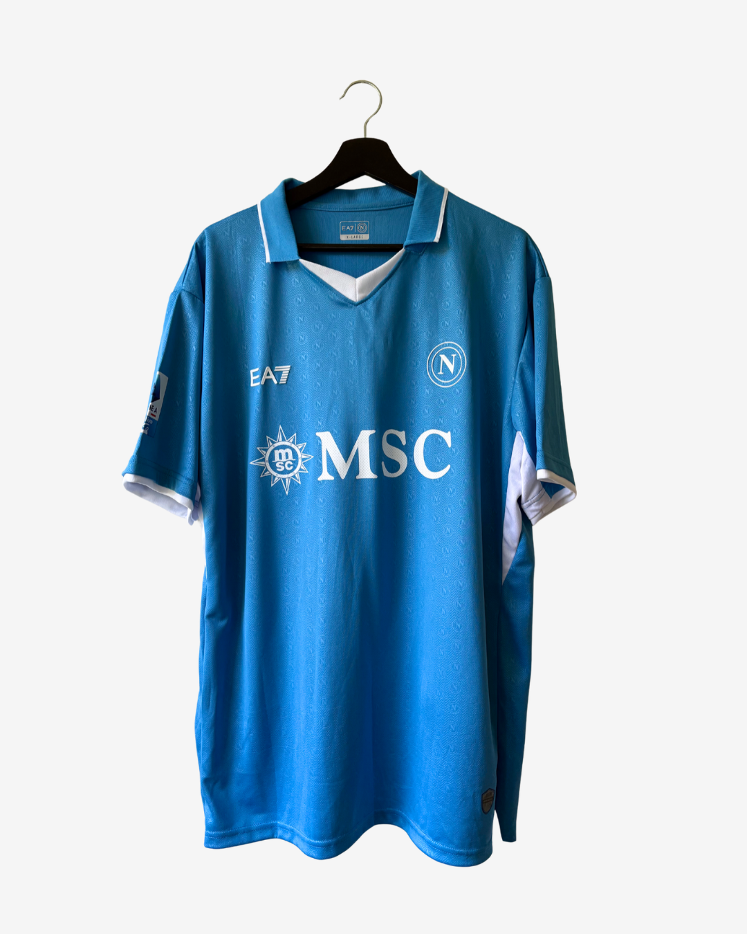 A blue vintage SSC Napoli soccer jersey hanging on a black hanger against a light gray background. The jersey features white trim on the collar and cuffs, the white 'MSC' sponsor logo on the chest, the SSC Napoli crest, and the year '1987' printed on the right chest.