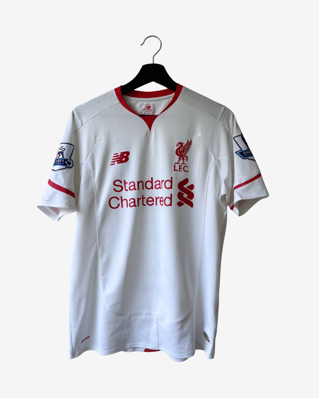 A white Liverpool football jersey with red trim, featuring the red New Balance logo, Liverpool FC crest, and Standard Chartered sponsor logo, hanging on a black hanger against a light gray background.