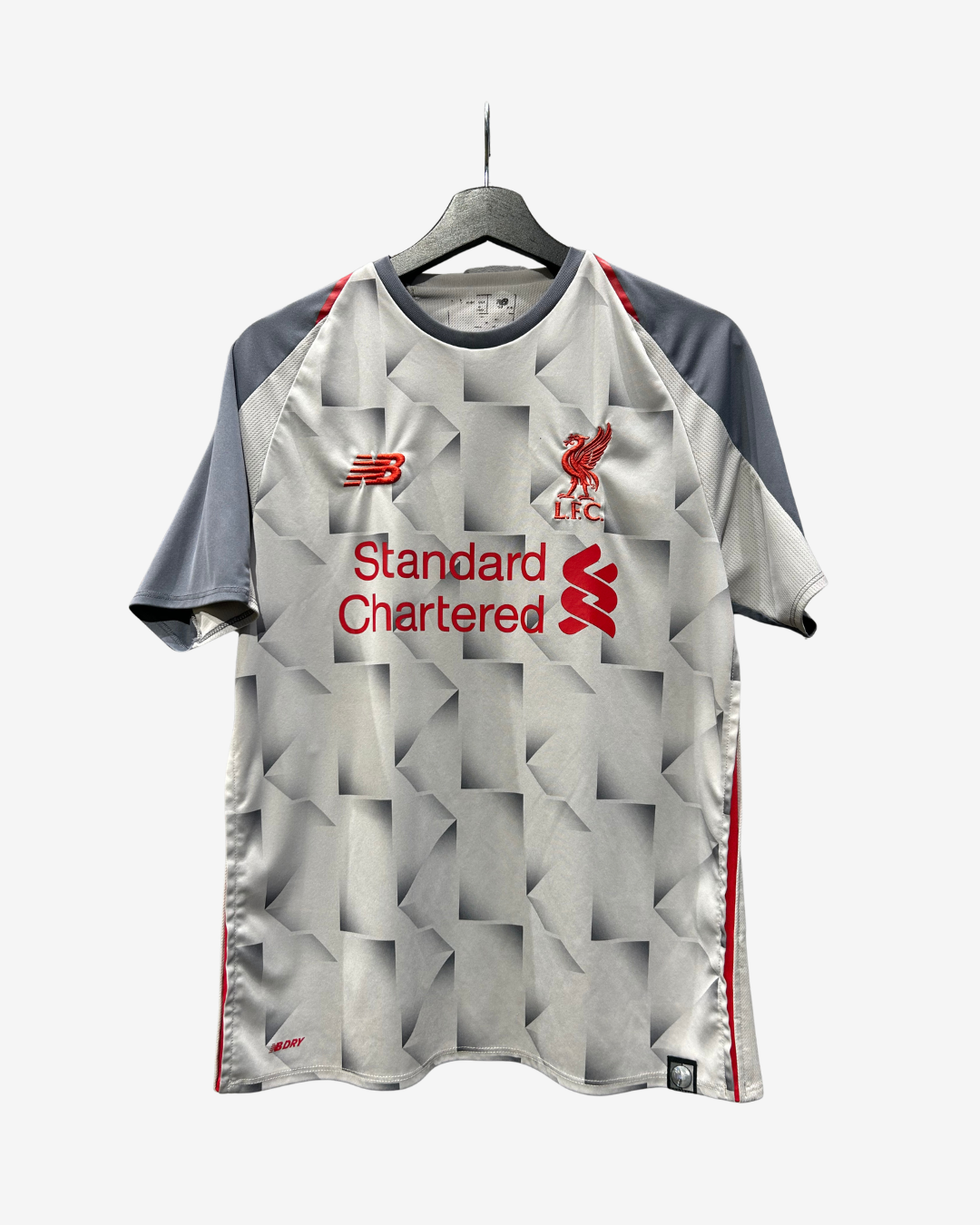 A short-sleeve Liverpool football jersey hanging on a black hanger against a light gray background. The jersey features a silver-gray geometric pattern with red accents, including the red Liverpool FC crest, New Balance logo, and Standard Chartered sponsor logo.