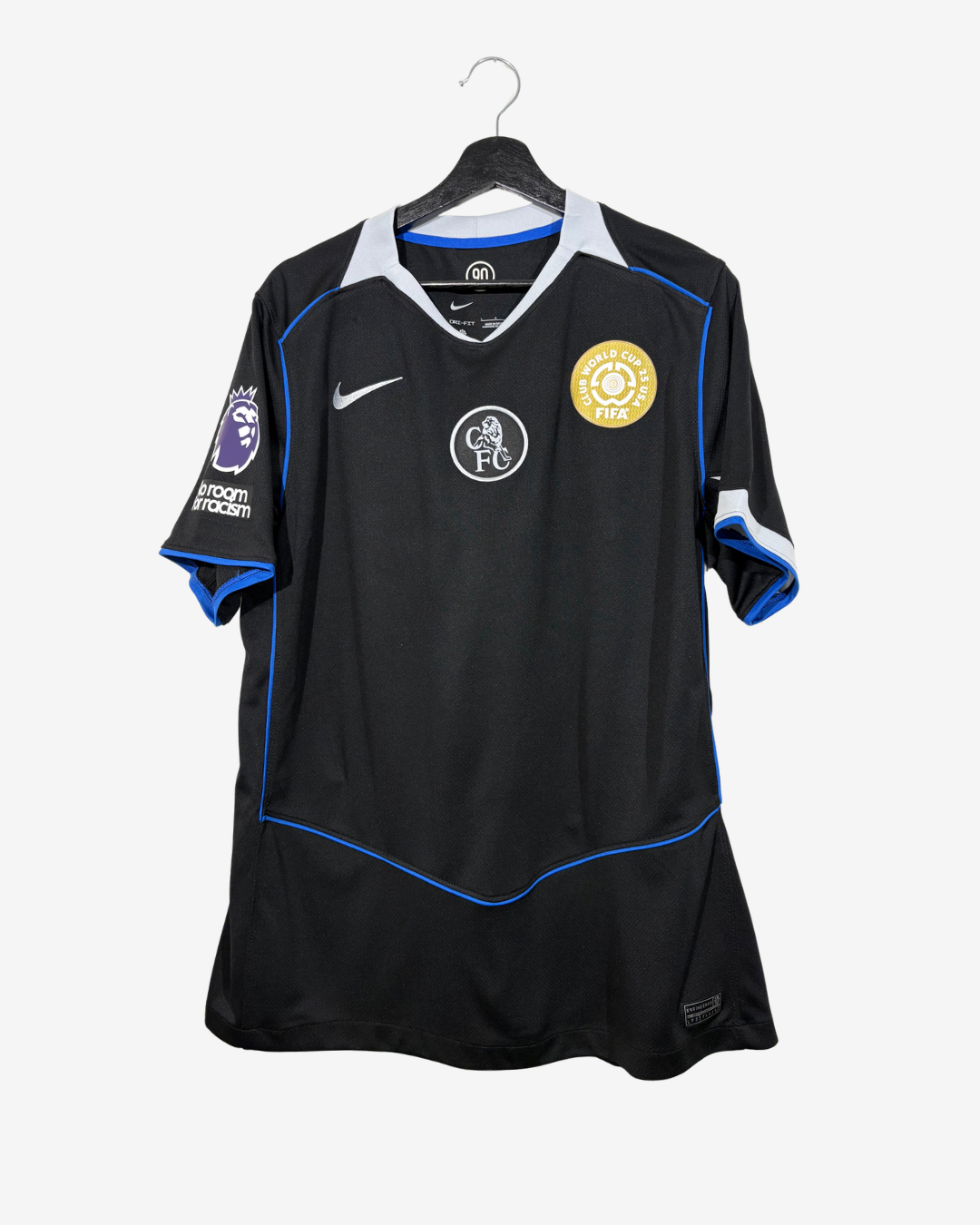 A black Chelsea FC Nike soccer jersey with blue and white accents, hanging on a black hanger against a light gray background. The jersey features the club crest on the left chest, a Nike swoosh on the right chest, and a Premier League patch on the sleeve.