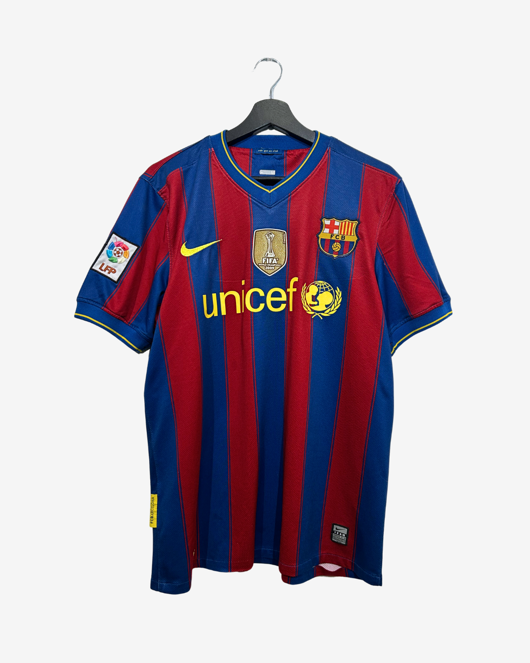 A vintage FC Barcelona home soccer jersey with red and blue vertical stripes, featuring the yellow Nike swoosh, unicef sponsor logo, and LFP patch on the sleeve, hanging on a black hanger against a light gray background.