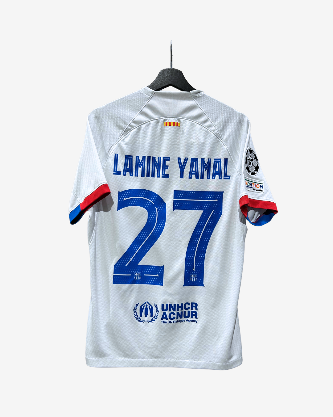 White soccer jersey with 'LAMINE YAMAL' and the number '27' in blue on the back, hanging on a black hanger against a light gray background.