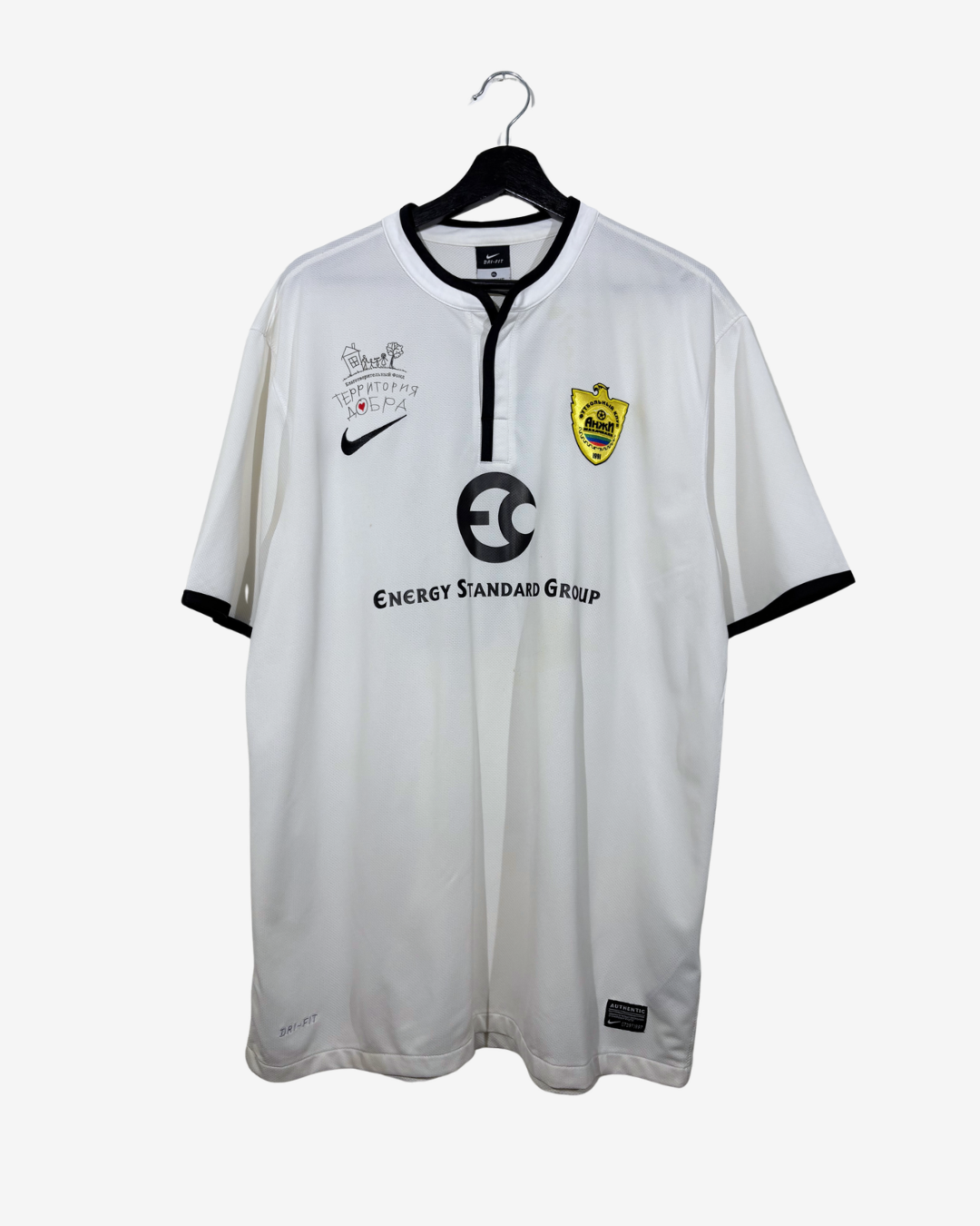 A white Anzhi Makhachkala soccer jersey with black trim, featuring the Nike swoosh, Energy Standard Group sponsor logo, and the team crest, hanging on a black hanger against a light gray background.
