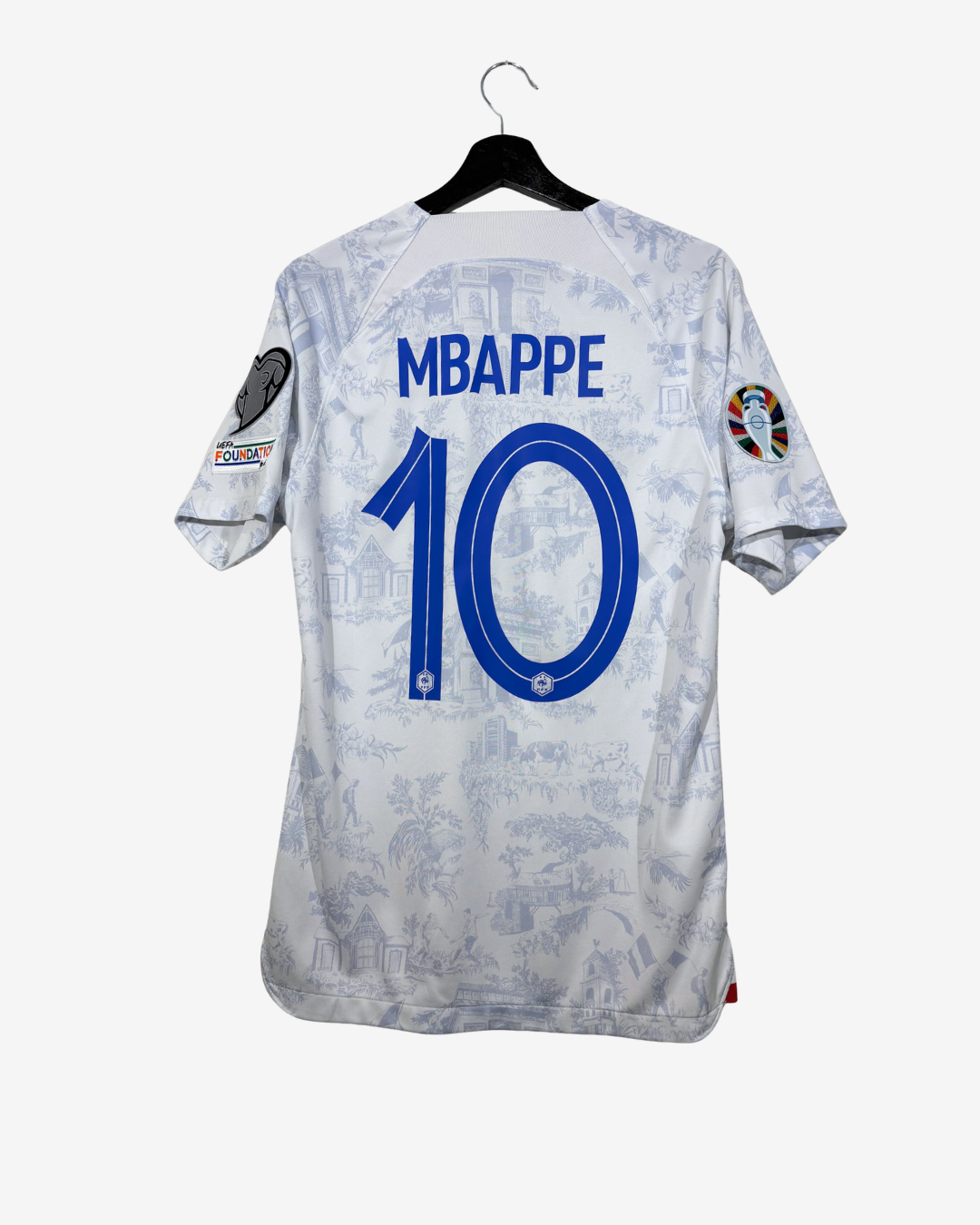 Back view of a white and blue patterned soccer jersey hanging on a black hanger against a light gray background. The jersey has 'MBAPPE' and the number '10' printed in blue on the back, with World Cup 2022 patches on the sleeves.