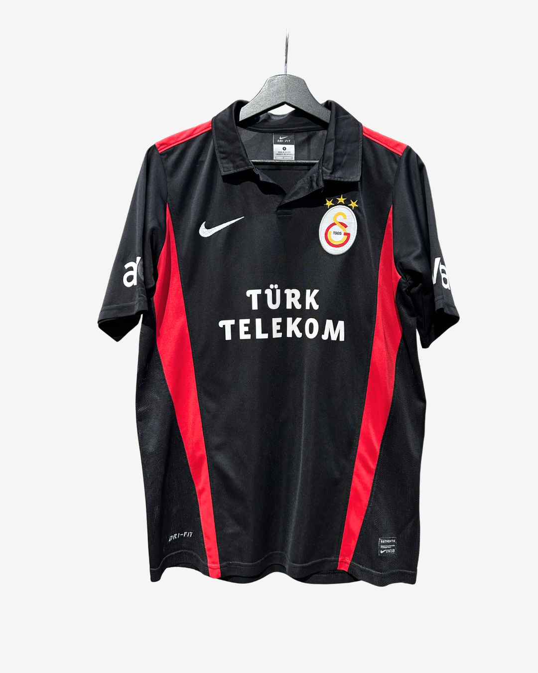 A black Nike Galatasaray sports jersey with red side panels and white logos, hanging on a gray hanger against a light gray background.