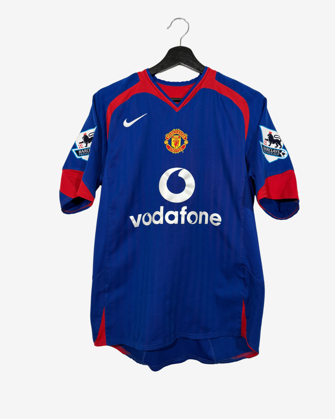 A vintage blue Manchester United soccer jersey hanging on a black hanger against a light gray background. The jersey features red shoulder panels, a white Nike swoosh, a Manchester United crest, and a white Vodafone sponsor logo.