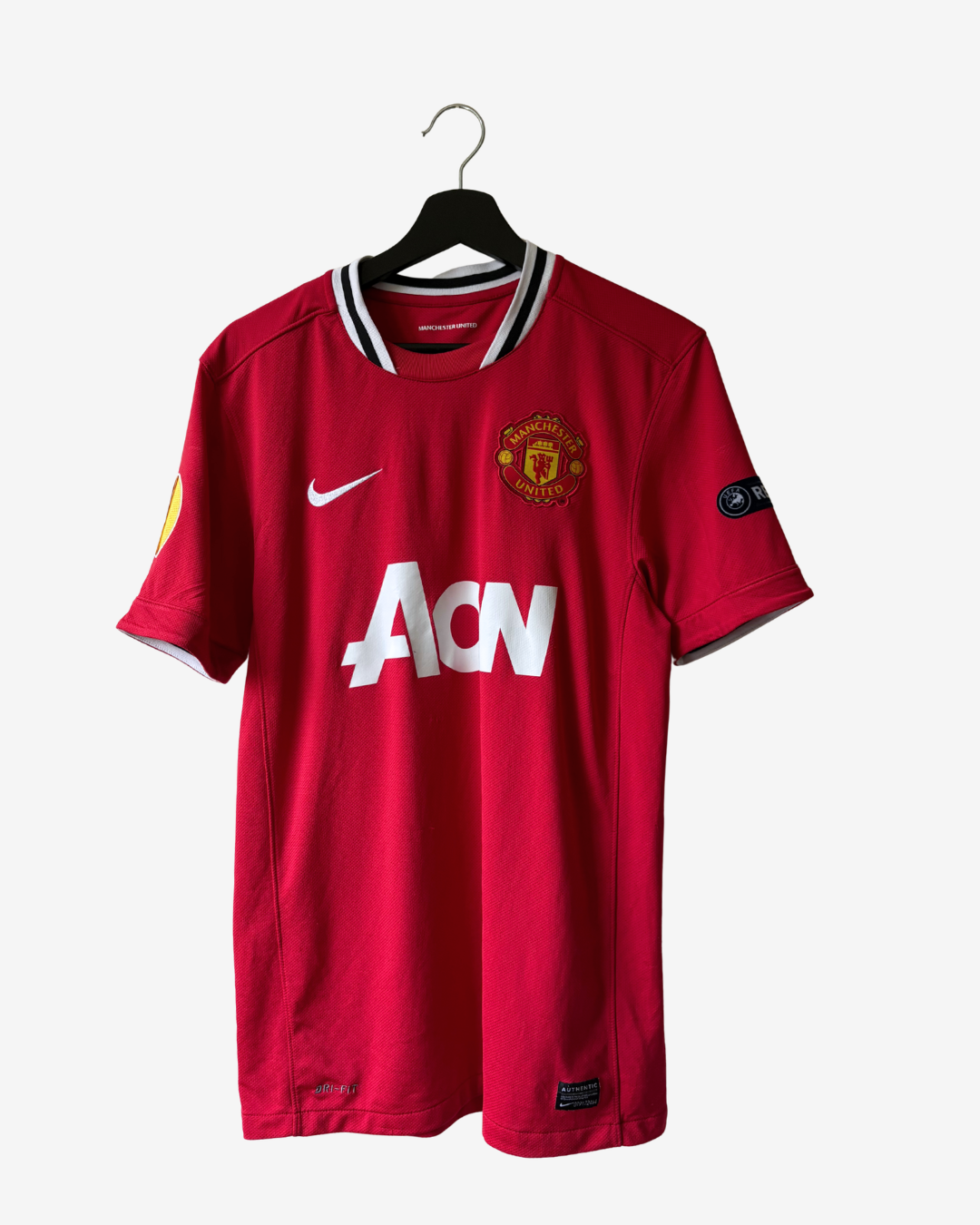 A red Manchester United Nike soccer jersey hanging on a black hanger against a light gray background. The jersey features the white 'AON' sponsor logo on the front, the Manchester United crest on the left chest, and a white Nike swoosh on the right chest.