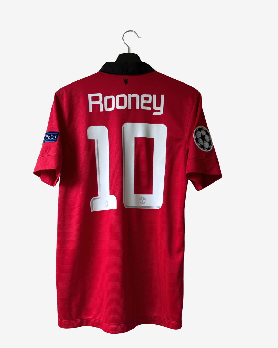 A red short-sleeve soccer jersey hanging on a black hanger against a light gray background. The back of the jersey shows the name 'Rooney' and the number '10' in white, with a UEFA Champions League patch on the sleeve.