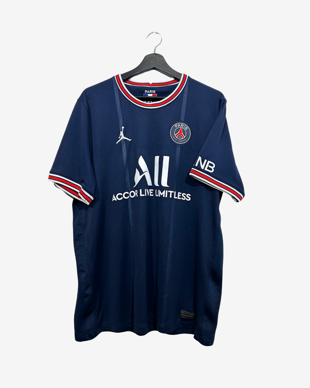 A Paris Saint-Germain Nike soccer jersey in navy blue, featuring the PSG crest, Jordan logo, and NB sponsor logo, hanging on a black hanger against a light gray background.