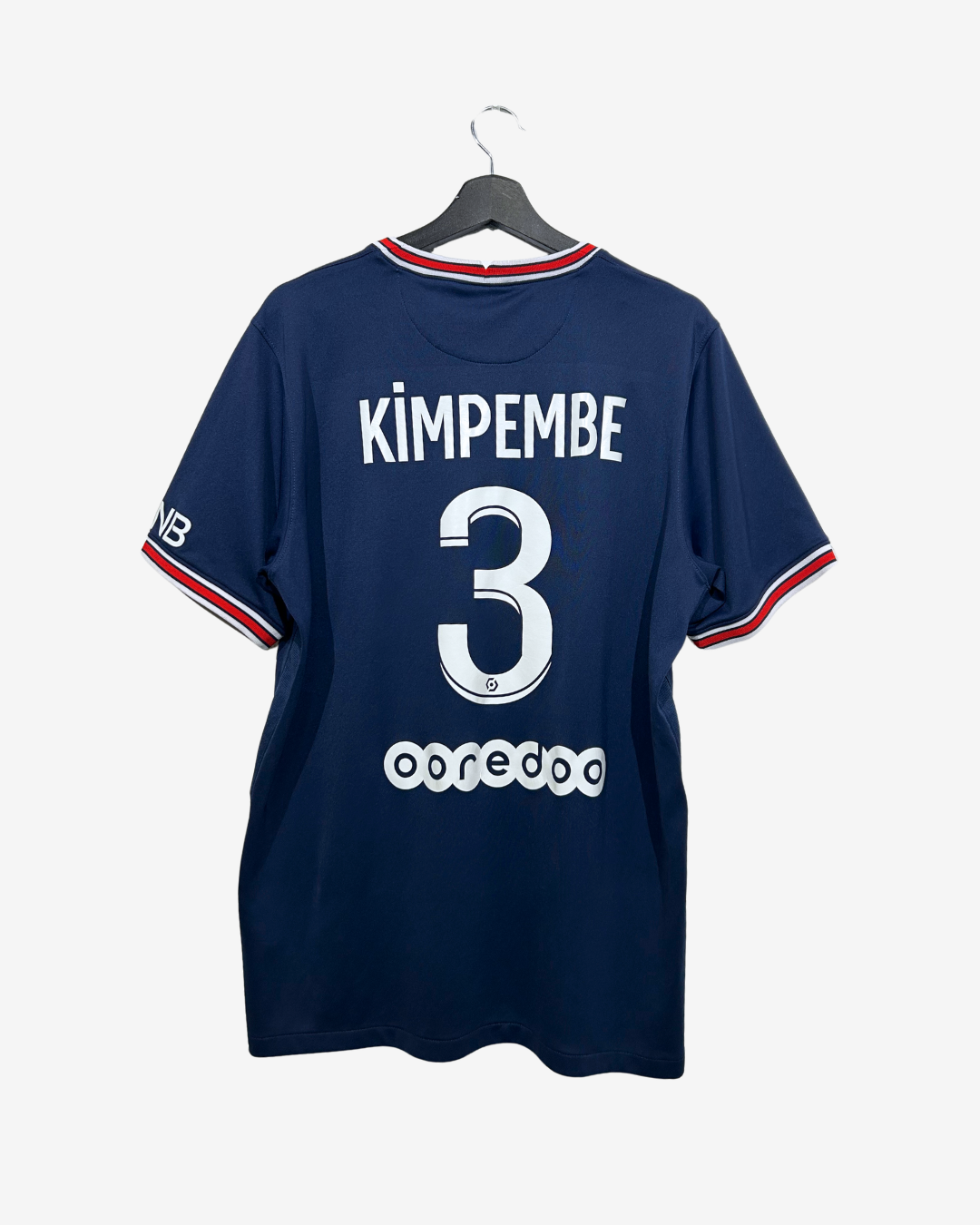A navy blue short-sleeve sports jersey hanging on a black hanger against a light gray background. The back of the shirt features the name 'KIMPEMBE' and number '3' in white, with red and white striped trim on the collar and sleeve cuffs.