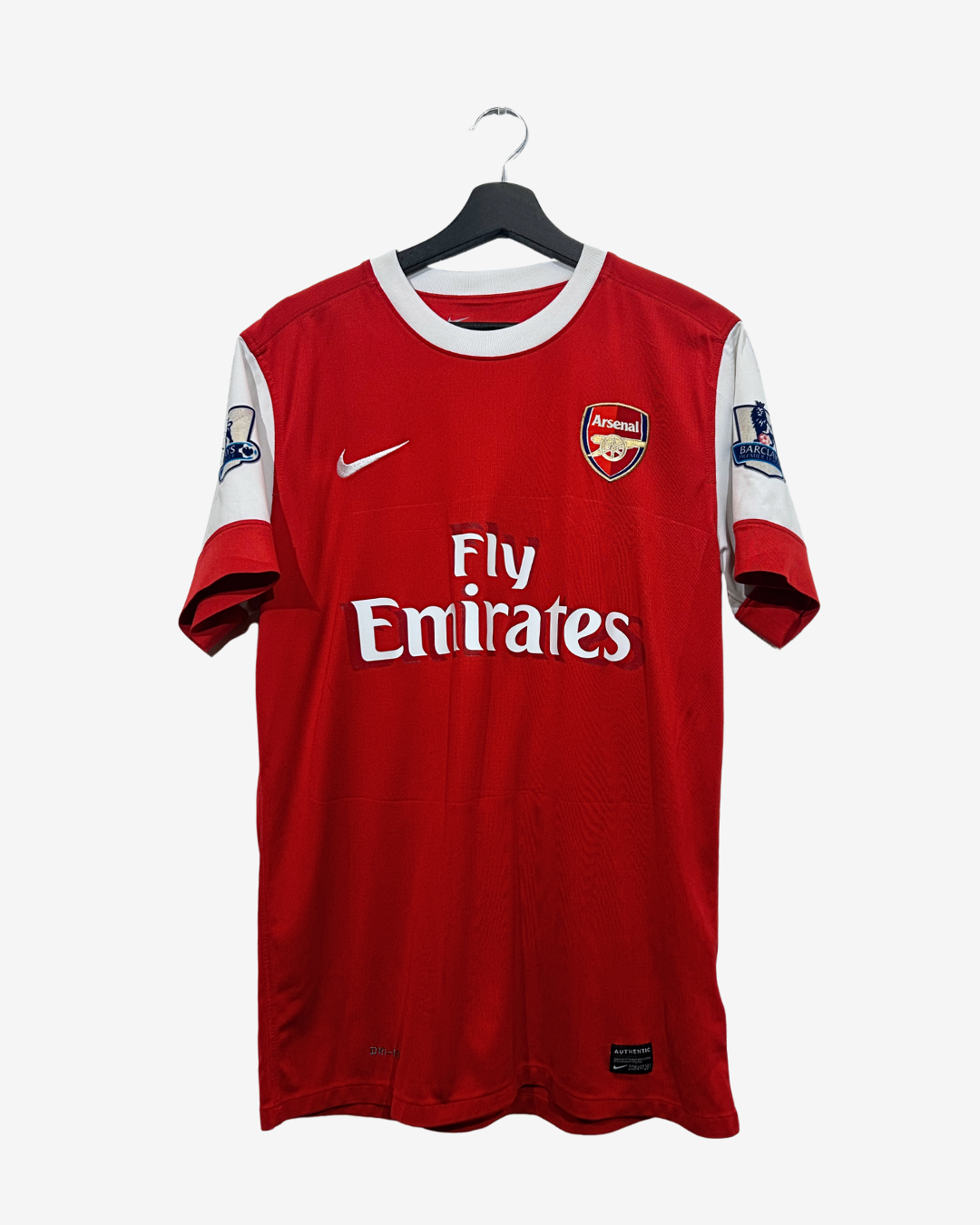 A red Arsenal soccer jersey with white sleeves, featuring the Fly Emirates sponsor logo and Nike swoosh, hanging on a black hanger against a light gray background.