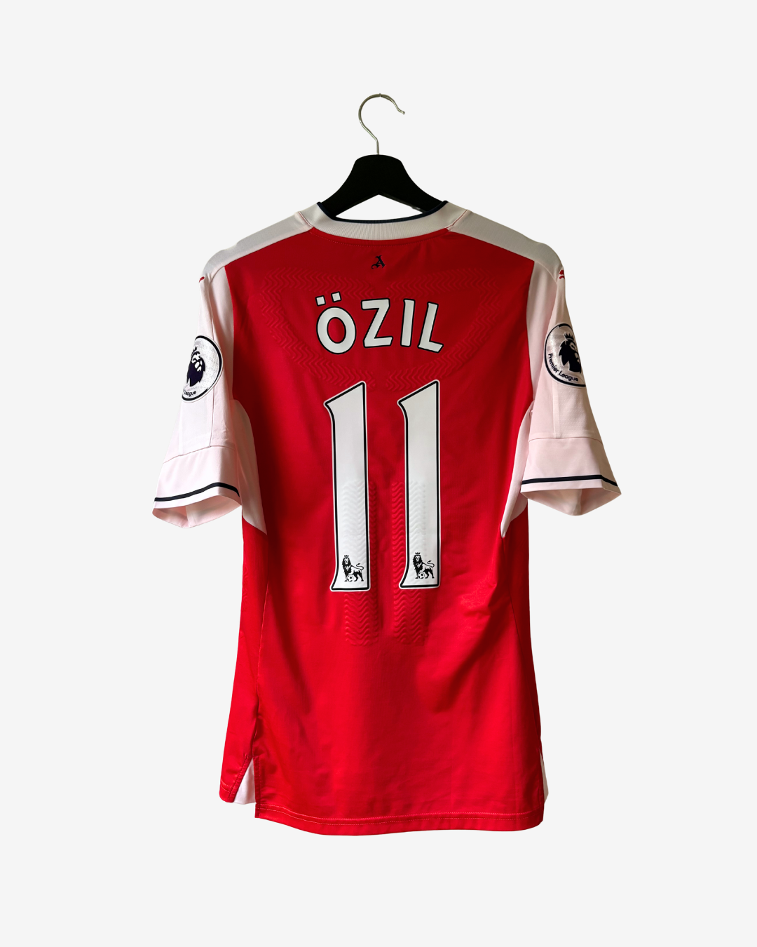 A red and white football jersey hanging on a black hanger against a light gray background. The back of the shirt features the name 'ÖZIL' and the number '11' in white, with Premier League patches visible on the sleeves.
