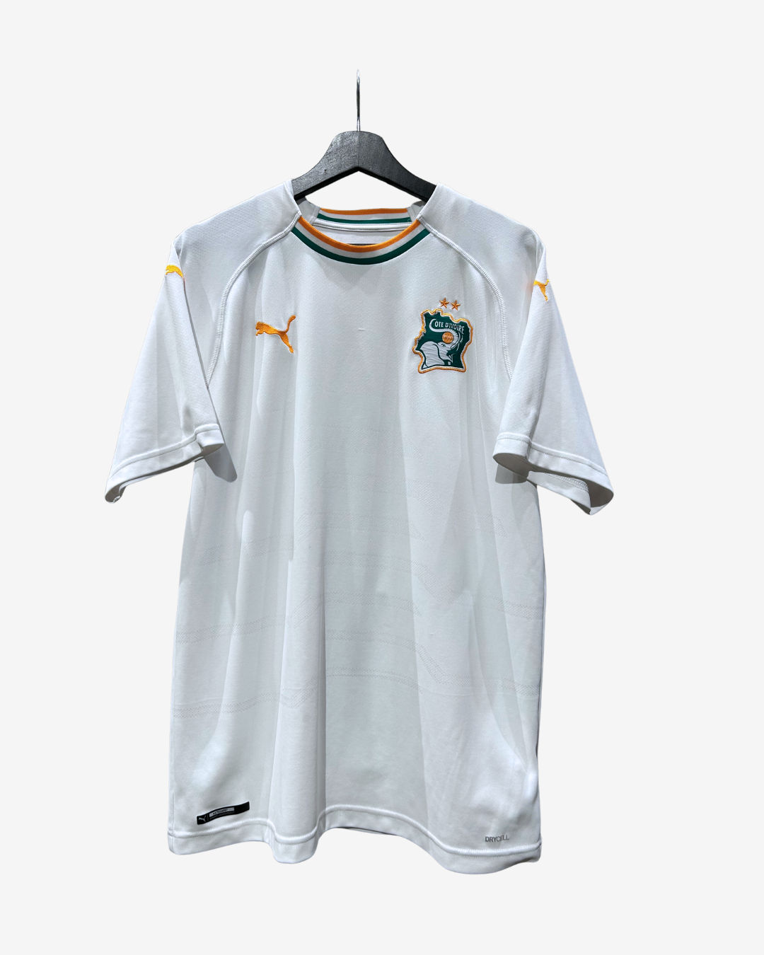 A white Puma Australia national football team home jersey hanging on a black hanger against a light gray background. The jersey features the yellow Puma cat logo on the right chest, the Australian Football League crest on the left chest, and a green, white, and orange striped collar.