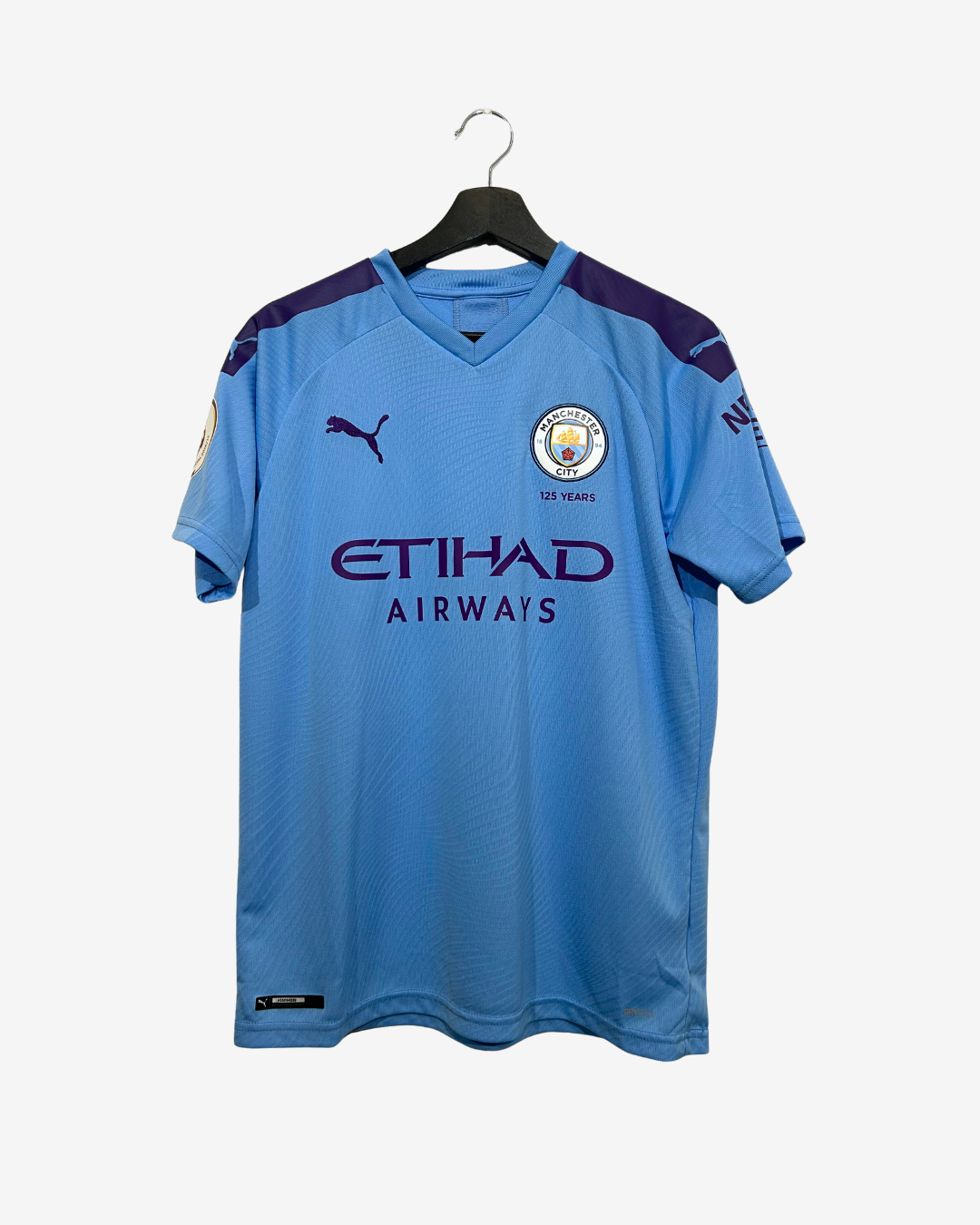 A light blue Manchester City soccer jersey by Puma, featuring the Etihad Airways sponsor logo, hanging on a black hanger against a light gray background.
