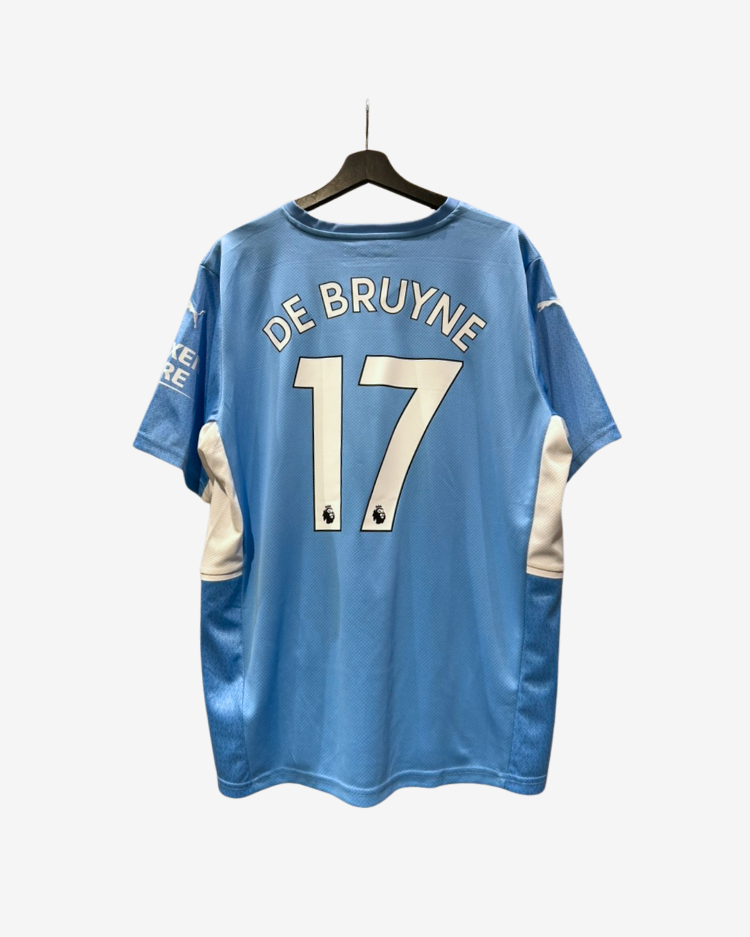 A light blue Manchester City soccer jersey with 'DE BRUYNE' and the number '17' printed in white on the back, hanging on a black hanger against a light gray background.