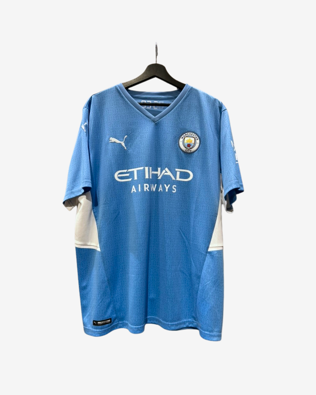 A light blue Manchester City soccer jersey by Puma, featuring the Etihad Airways sponsor logo on the front, hanging on a black hanger against a light gray background.