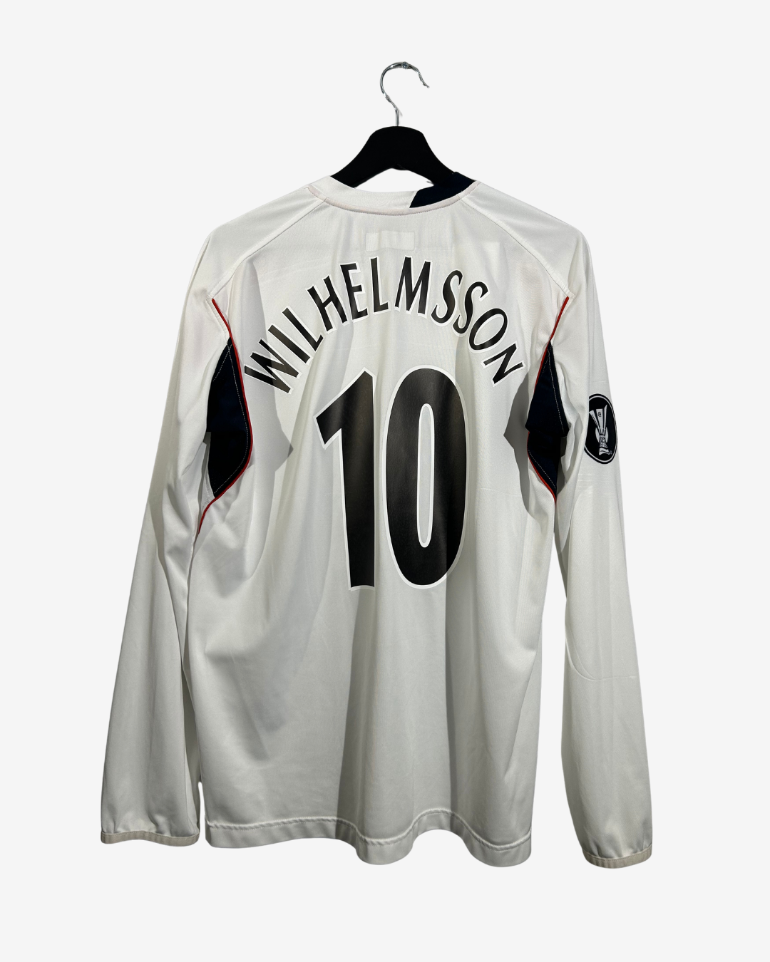 A white long-sleeve football jersey hanging on a black hanger against a light gray background. The back of the shirt features 'WILHELMSSON' and the number '10' in black, with black and red accents on the shoulders.