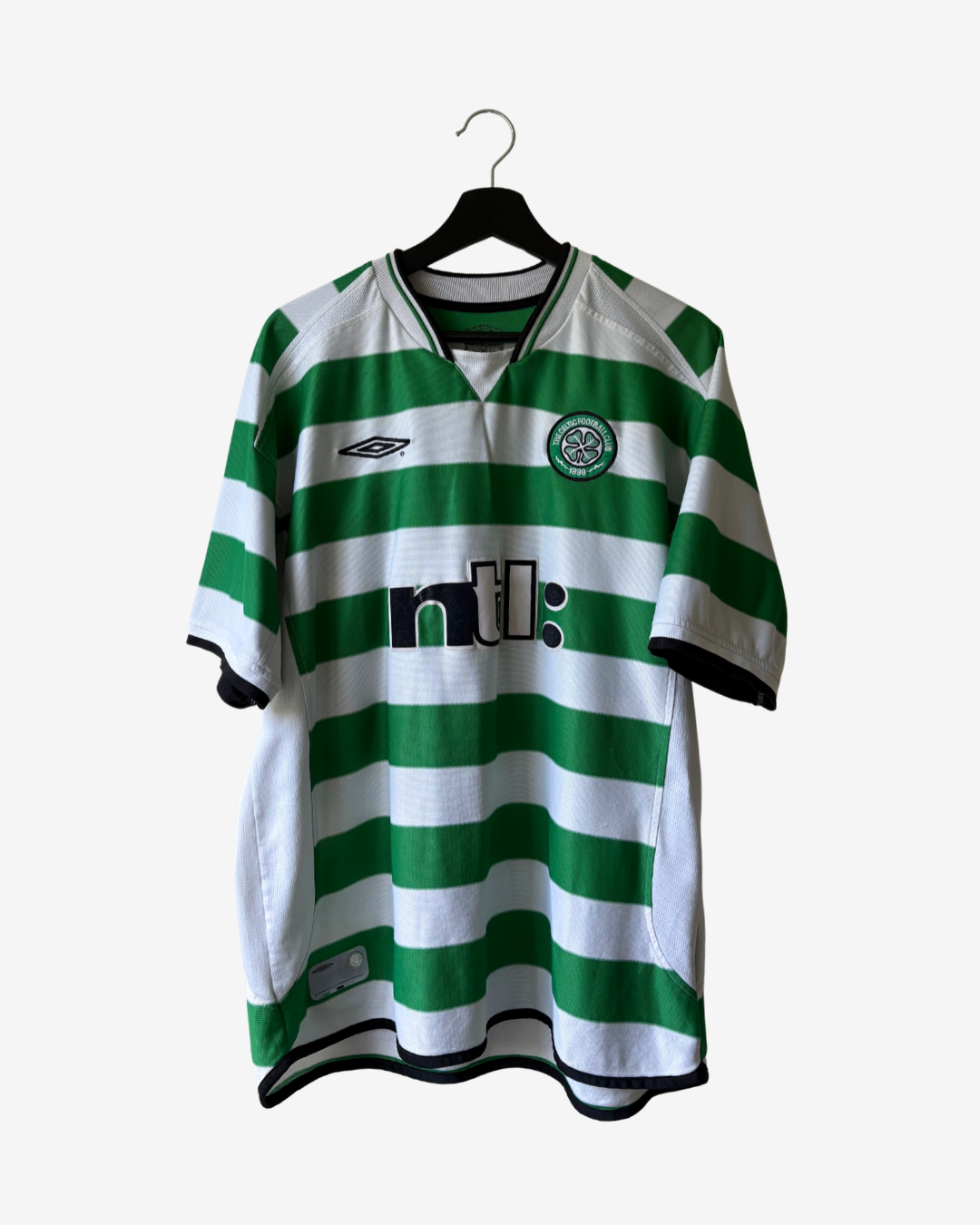 A vintage Celtic FC home soccer jersey by Umbro, featuring green and white horizontal stripes, hanging on a black hanger against a light gray background.