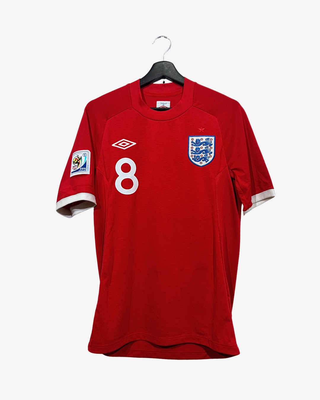 A red England national football team jersey by Umbro, featuring the number 8 on the front, the three lions crest, and a sleeve patch, hanging on a black hanger against a light gray background.